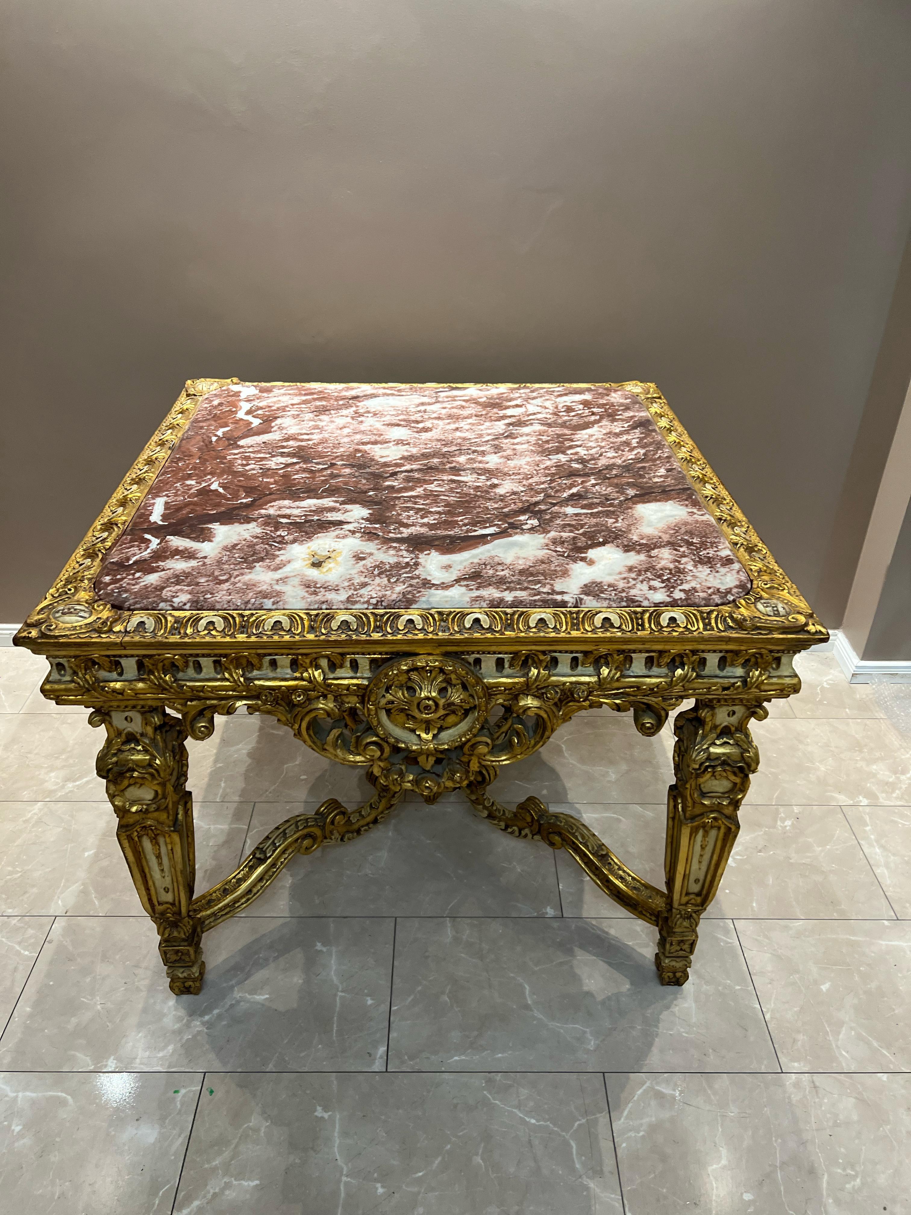 This is an ornate antique table with a luxurious and opulent design. The table features a square top made of polished marble with a striking blend of reddish-brown and white veining. The edges of the tabletop are framed with an elaborate gilded