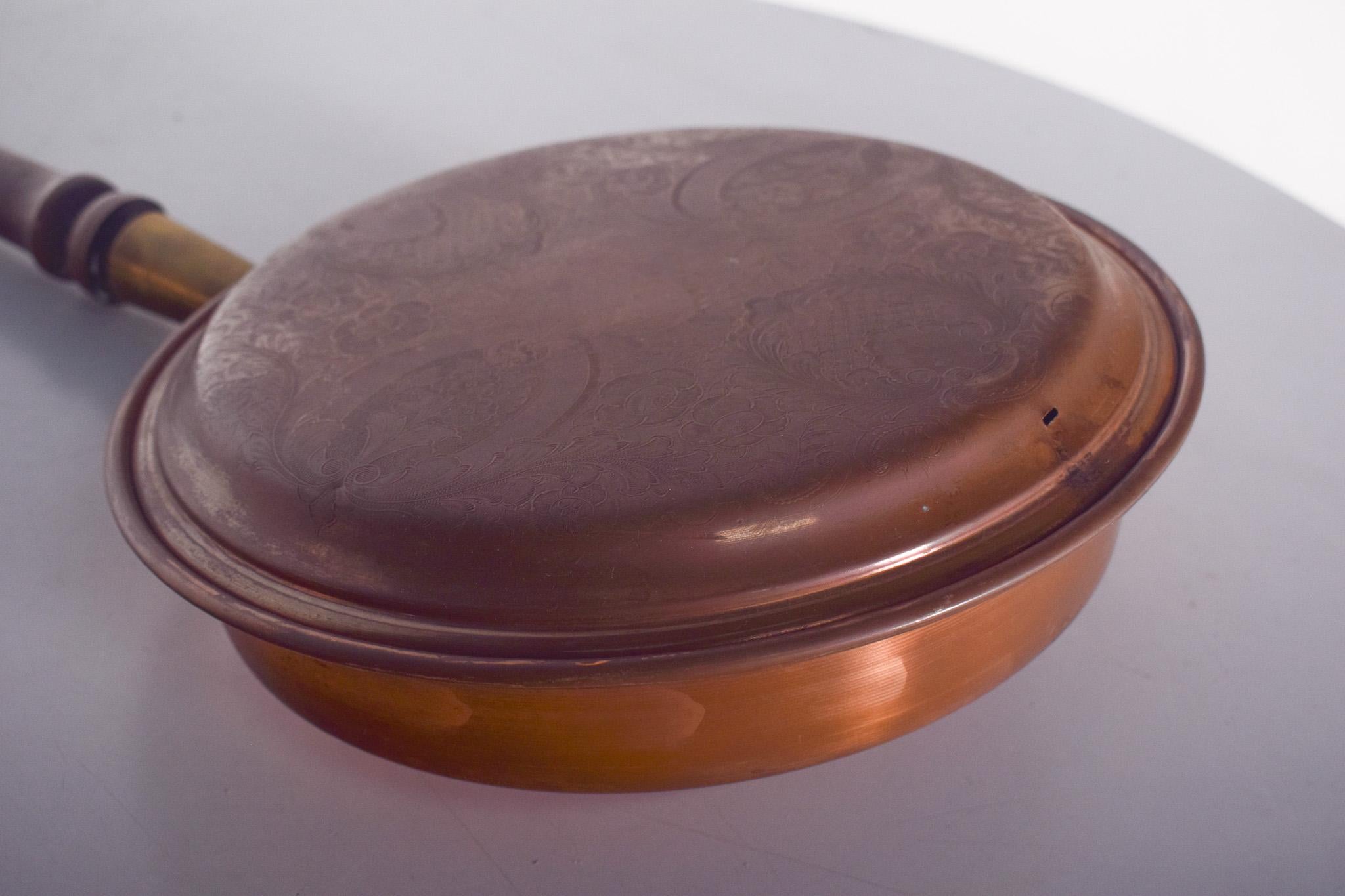 Brasero ancien avec poignée. L'objet consiste en un récipient en cuivre avec un couvercle décoré et de petits trous pour la ventilation. Le long manche, en bois tourné, permet une manipulation sûre sans risque de brûlure.
Pour fonctionner, des