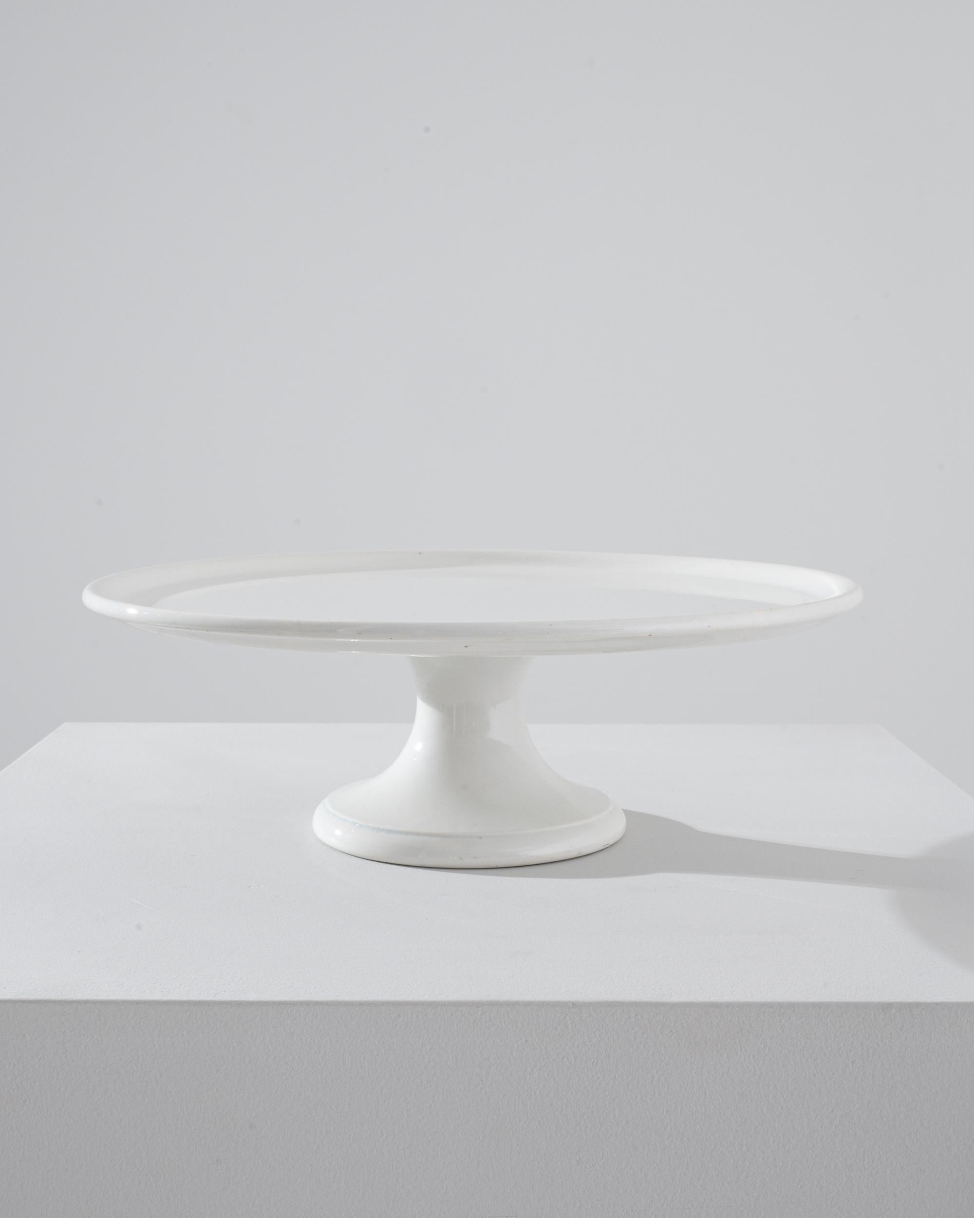 A ceramic cake tray from Belgium, produced circa 1900. A flat plane of circular ceramic coated in thick white glaze. With no unnecessary bells or whistles, this simple tray merely sits upon its stand and waits for cake. Only problem with this piece