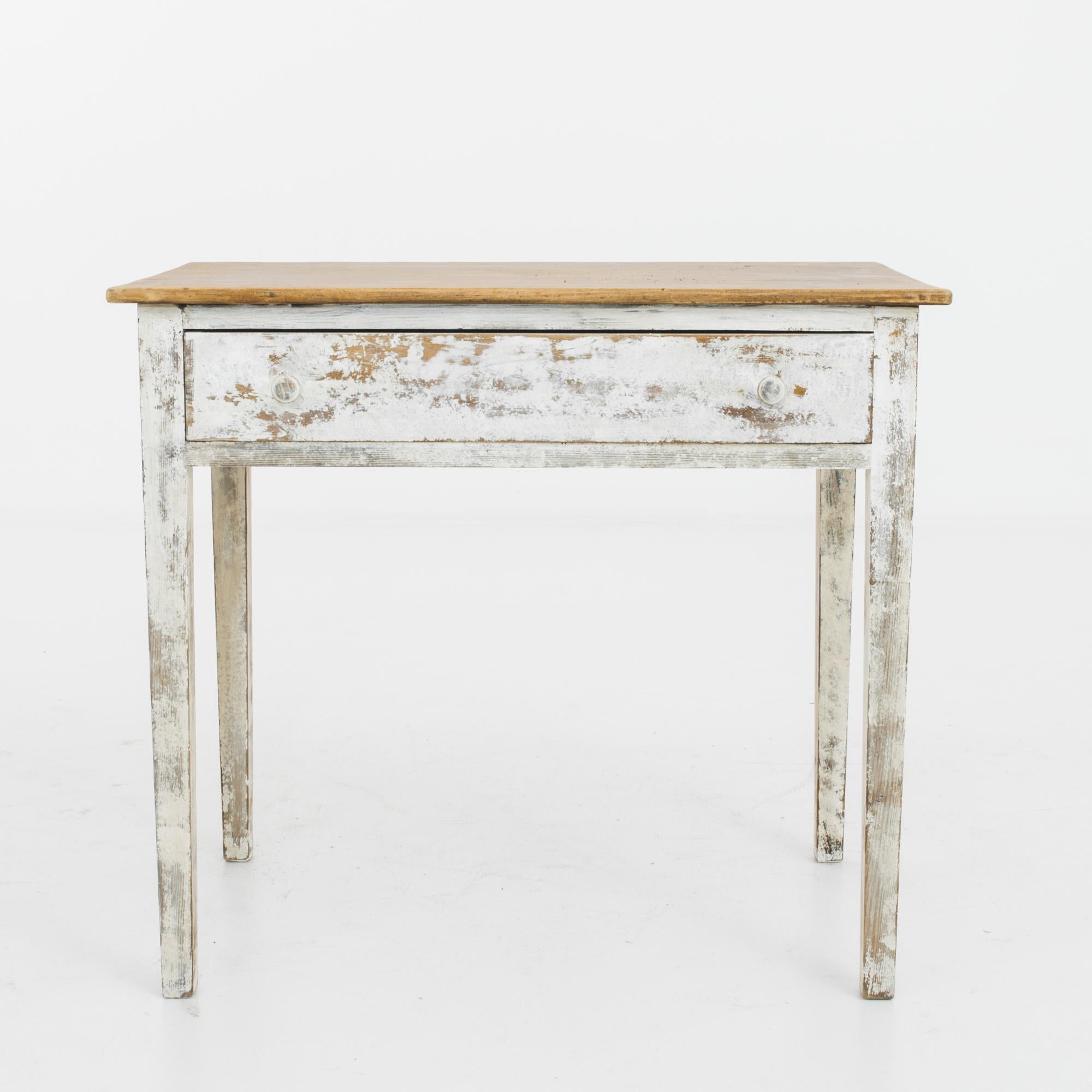 This antique wooden table was made in Belgium. It features a wide drawer with circular pulls and four legs that display a slight taper. A captivating rustic piece with its distressed white frame and the time-worn patina of the tabletop.