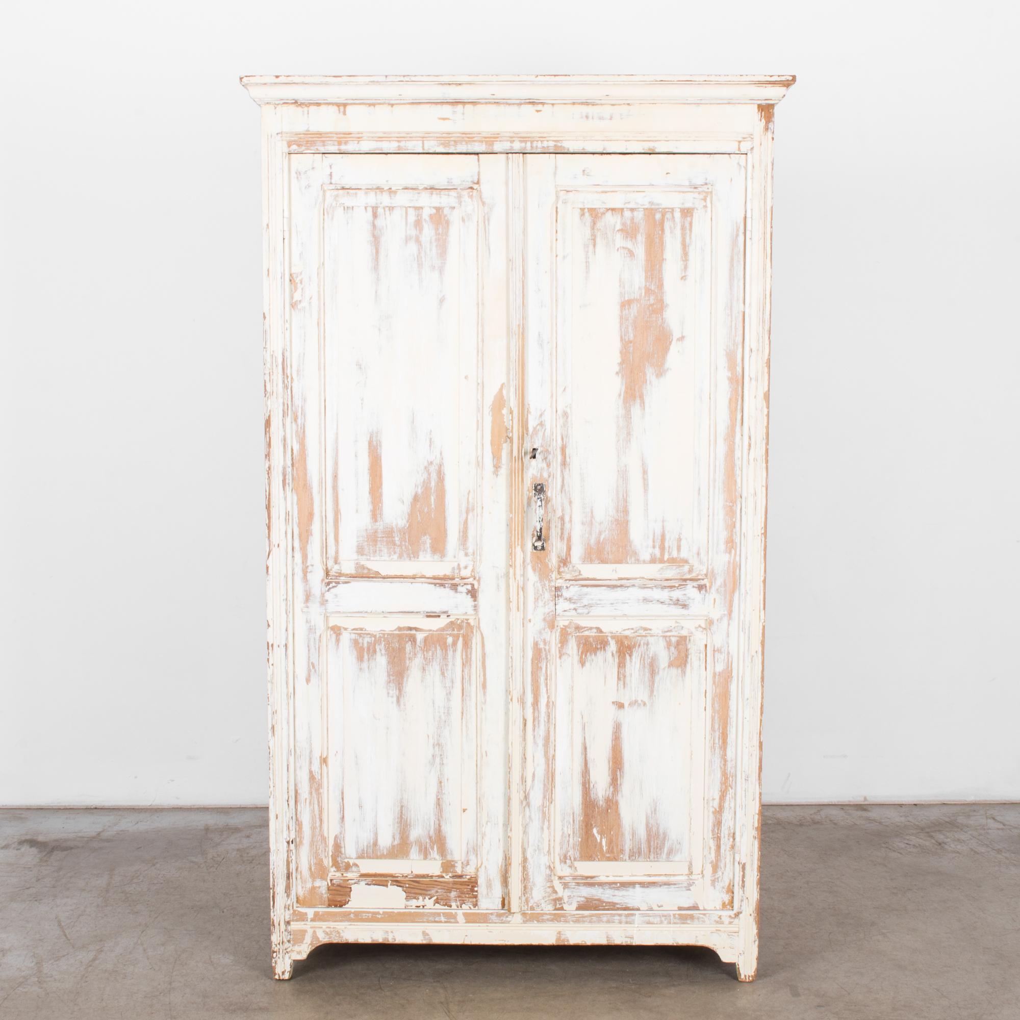 This wooden armoire with crown molding was made in Belgium. Originally painted white, the armoire displays a time-worn patina, which reveals the golden hues of the wood beneath and evokes the rustic charm of the countryside. The armoire is slightly