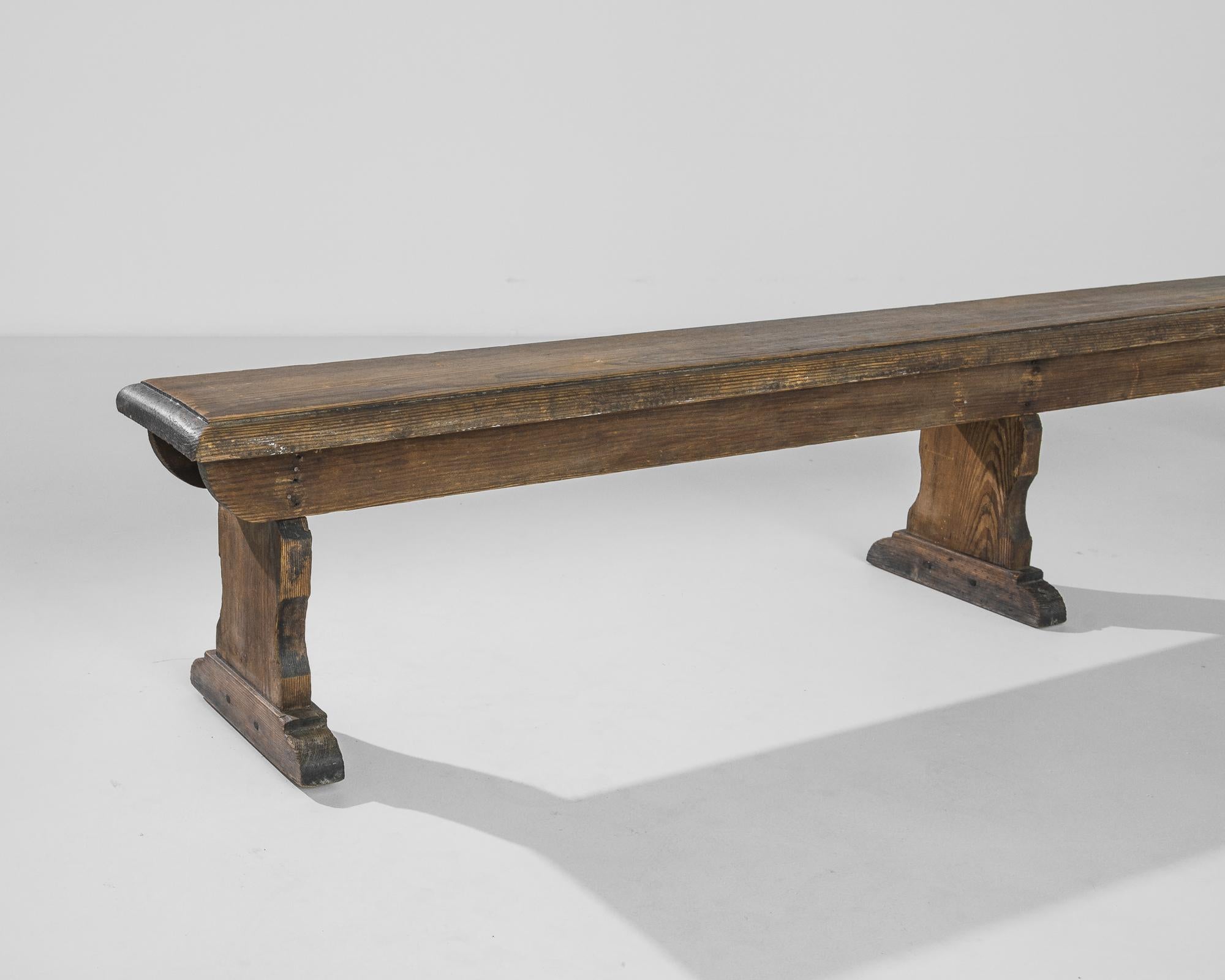 A wooden bench from the turn of the century Belgium. A long, slender bench sits atop a trio of trestle legs: the simplicity of the shape is reminiscent of a church pew. The deep umber tone of the wood is enhanced by a subtle polish, emphasizing the