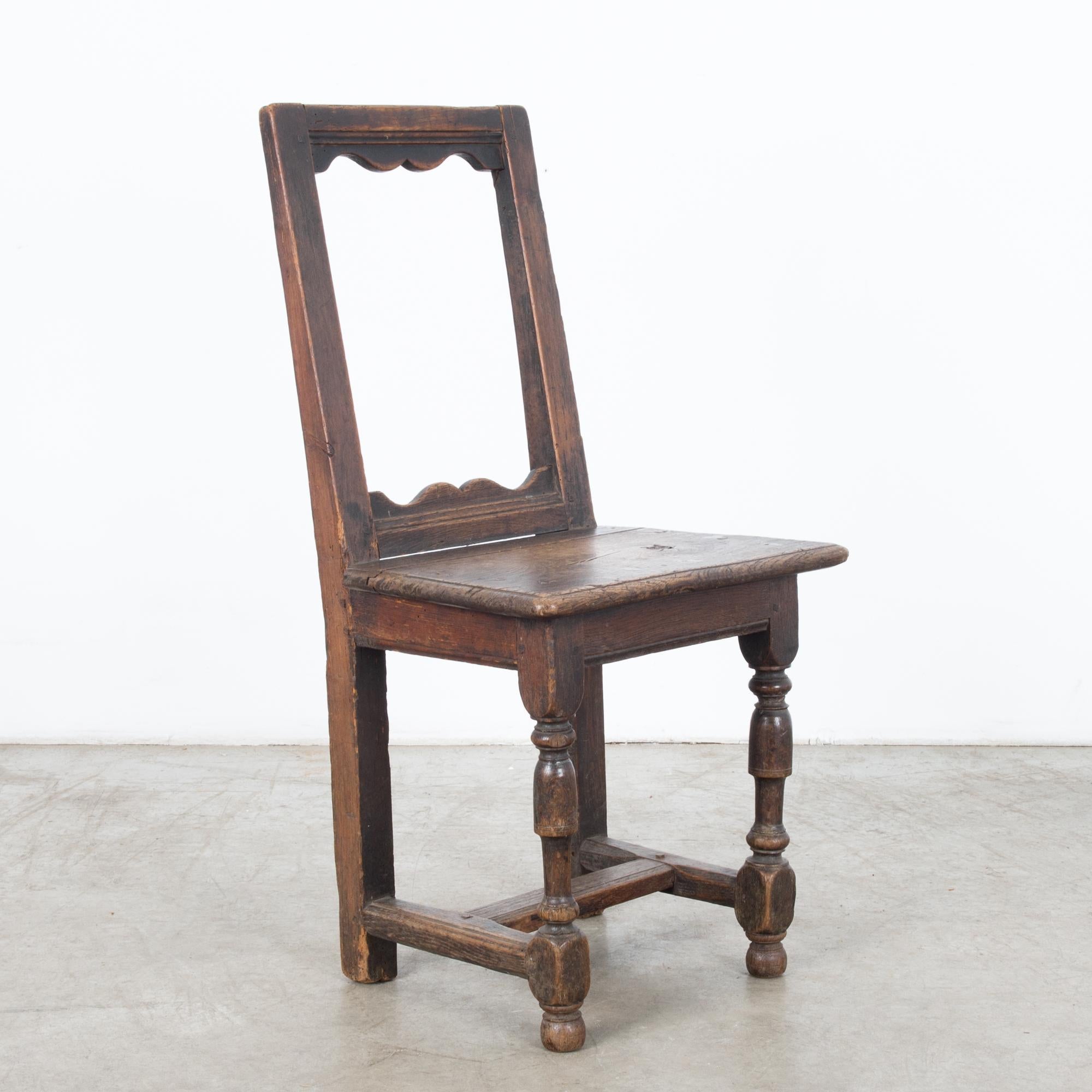 A wooden chair from Belgium, circa 1800. An upright shape with a striking open-framed backrest and a square paneled seat upon turned legs. The dark, softly polished wood has weathered into a patina with areas of highlight and shadow. The form of the