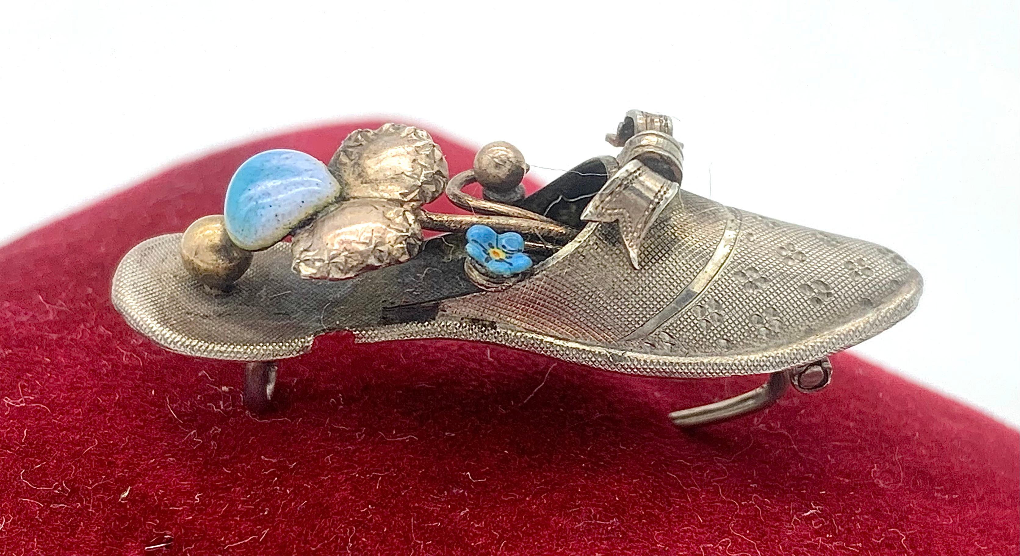 This beautiful and poetic brooch in the shape of a silver slipper is decorated with a bow and filled with enamelled flowers and a gilt ball. The shoe is finely engraved and marked 800 silver.