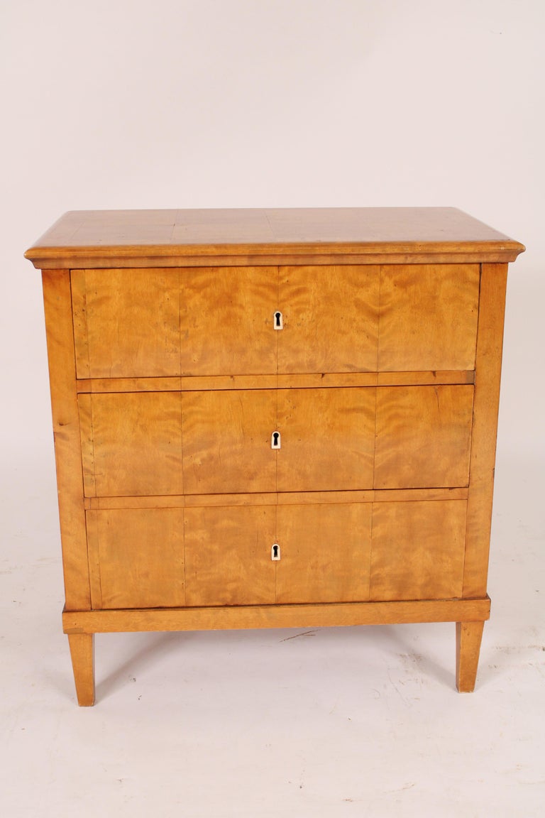 Antique Biedermeier style birch, ash and pine chest of drawers, 19th century. With an ash parquetry over hanging top over 3 birch drawers with bone escutcheons, pine sides resting on square tapered legs.