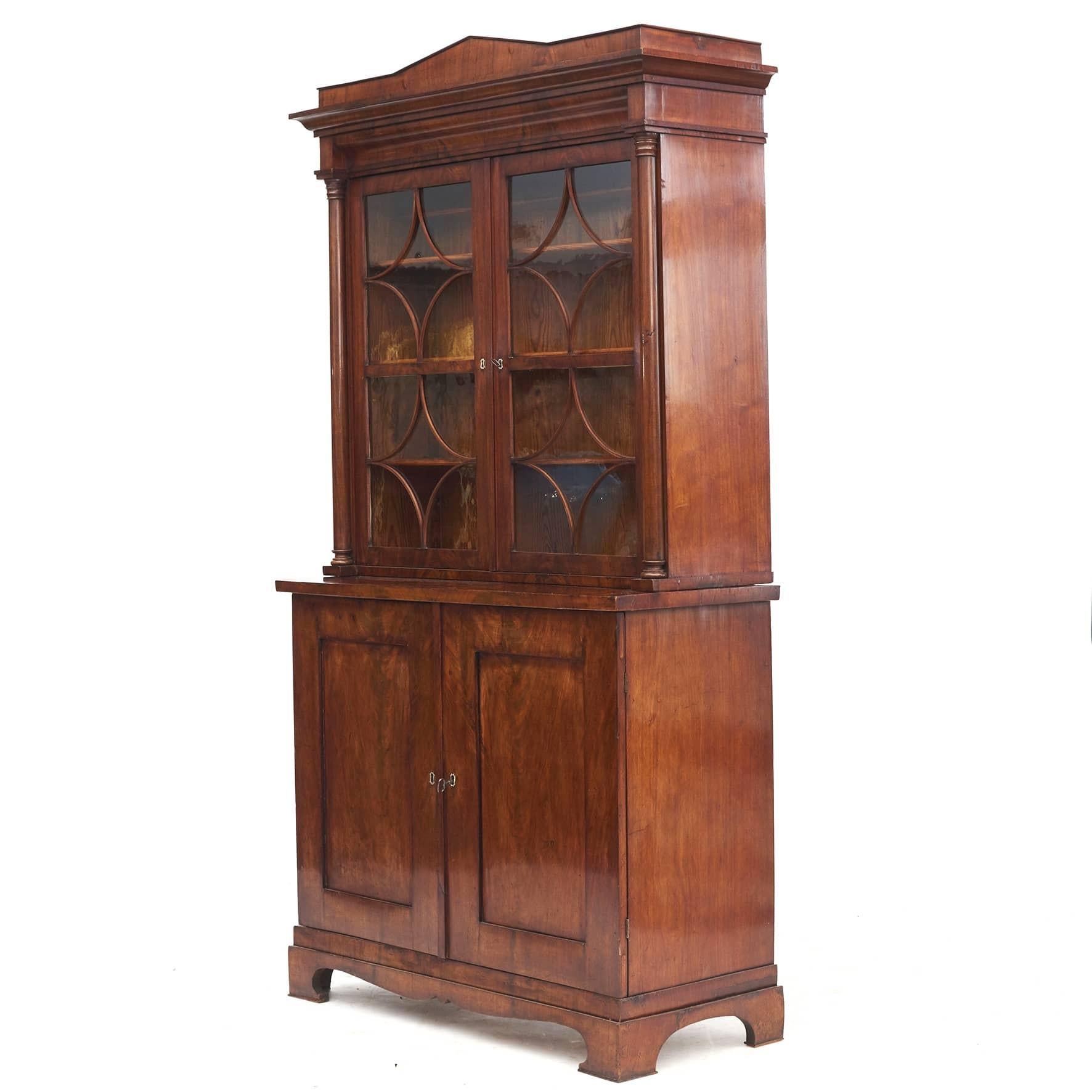 Biedermeier bookcase in mirror-cut flamed mahogany. Neoclassical style.
In 2 parts: Wall cabinet with architectural top. Pair of glass doors and concave bars with lemon marquetry, flanked by free-standing columns.
Base cabinet with a pair of doors