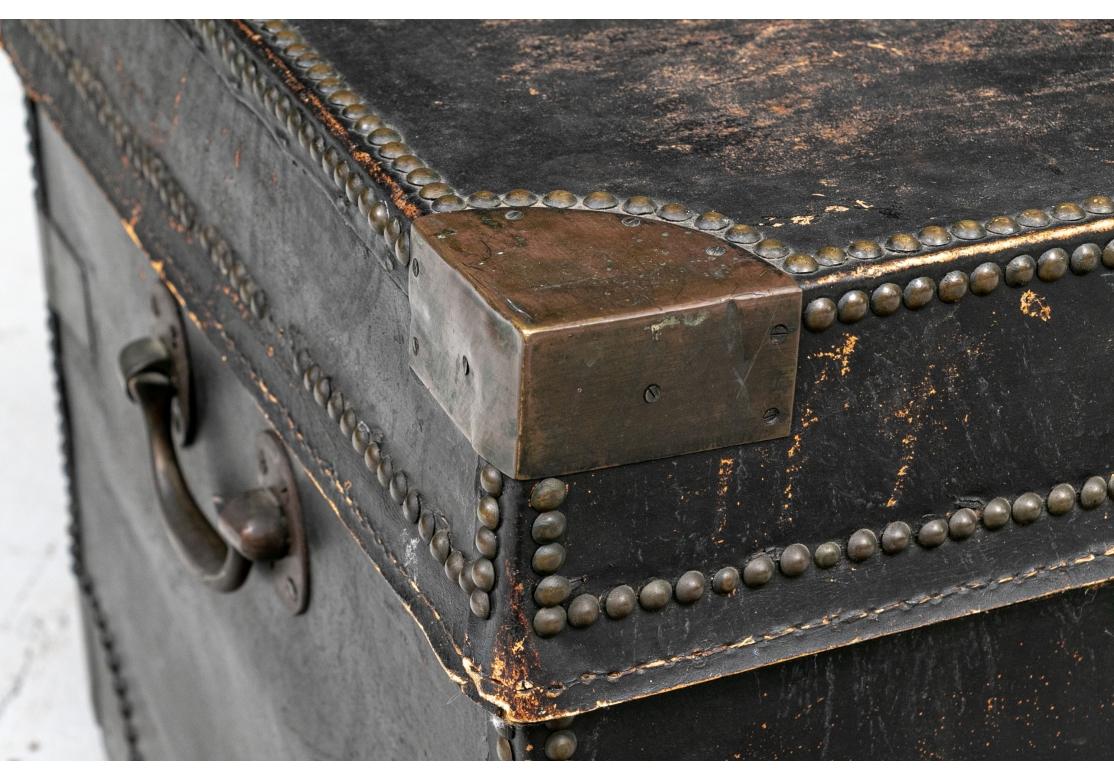 Antique black leather campaign trunk with all over brass nail head decoration including an oval medallion on lid. The trunk with brass corners, supports, shield form escutcheon, side handles, ring pull hardware and resting on a footed wood