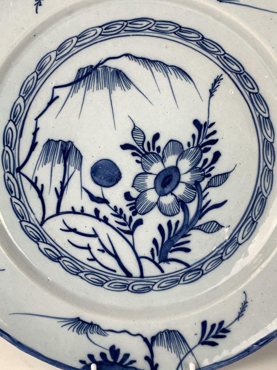 This lovely Delft charger was hand-painted in England in the mid-18th century, circa 1765.
The center shows a naive garden scene with a willow tree, an oversized flower, and a large blue bud.
The border decoration echoes the center scene.
It is