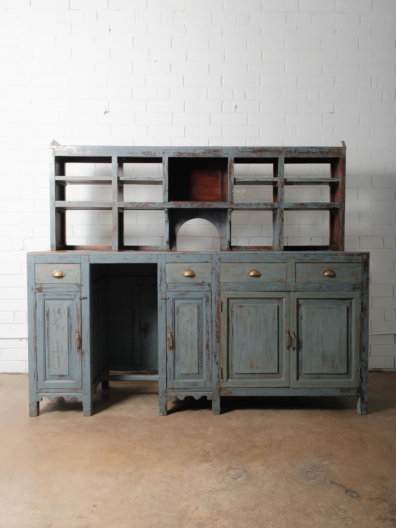 Un notevole bancone antico in legno verniciato di blu con una credenza superiore, caratterizzato da strati di vernice splendidamente consumati che rivelano decenni di utilizzo e di storia. Questo pezzo sostanzioso offre una rara combinazione di