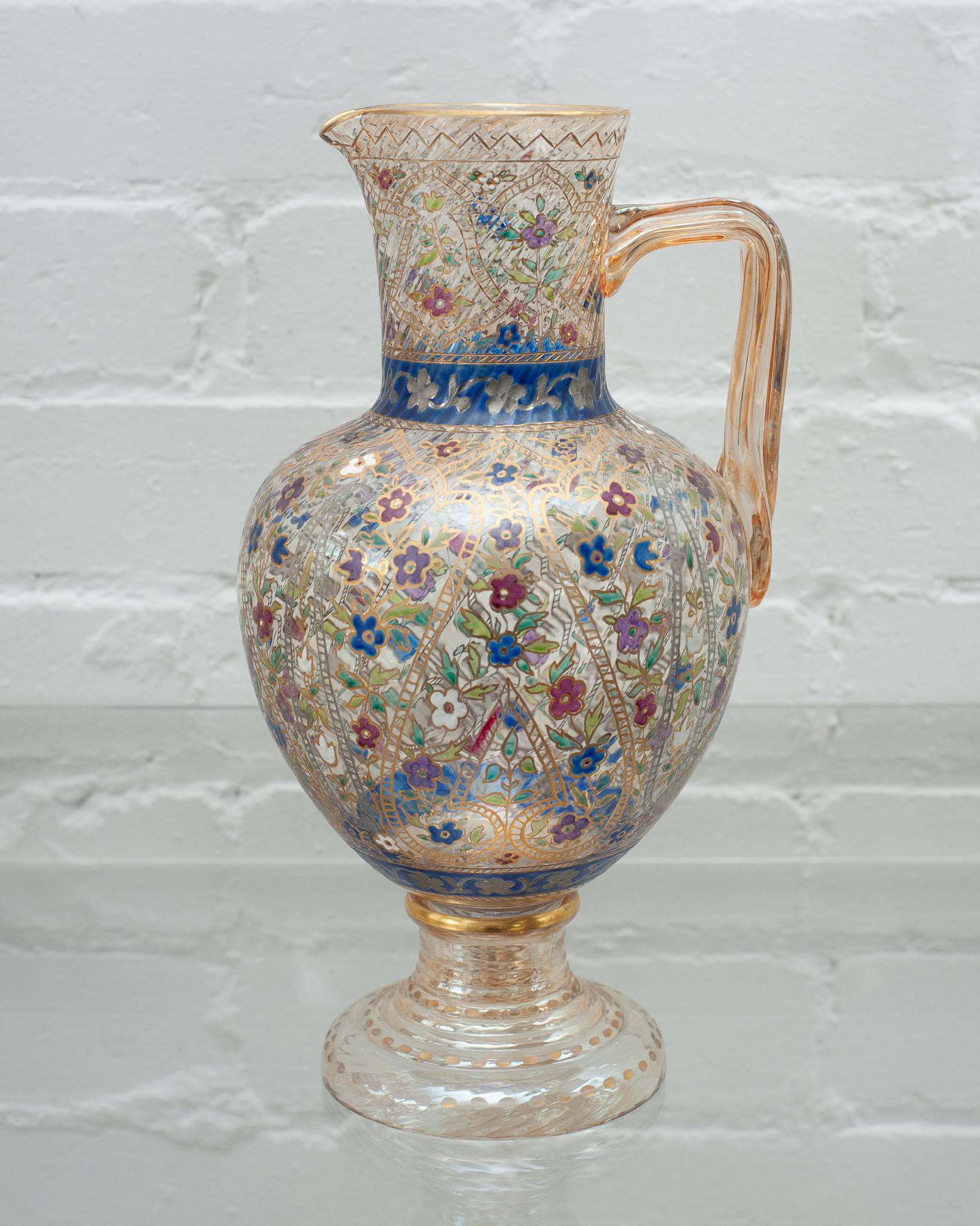 A gorgeous antique Bohemian glass water pitcher with ornate enamel work, circa 1920. Decorated in floral motifs and gold gilding, this vessel will brighten up any table with its fine craftsmanship and bold colour.