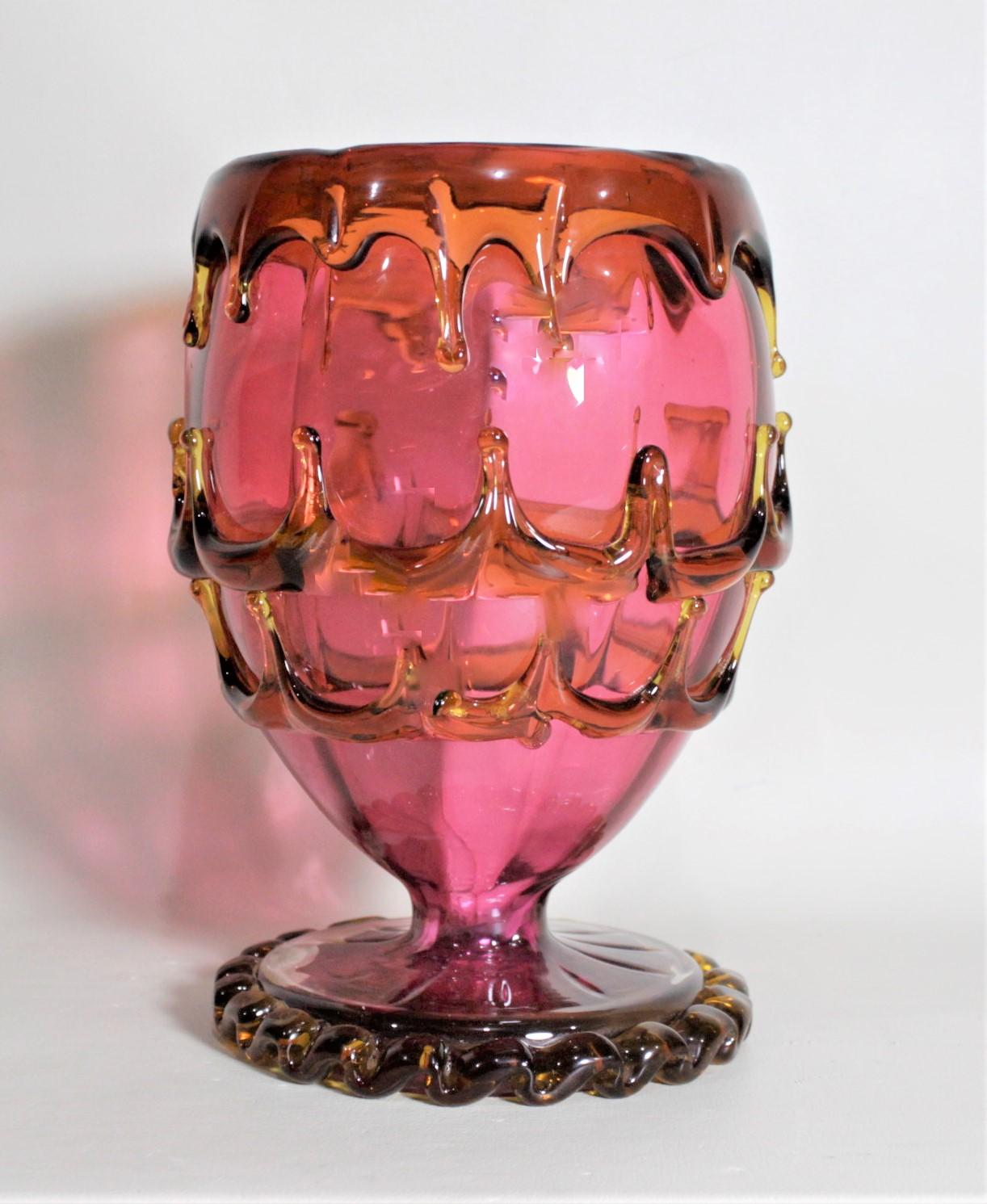 Cet ancien vase sur pied en forme de calice en canneberge n'est pas marqué mais on pense qu'il a été fabriqué en Bohême vers 1880. Ce vase sur pied est tout à fait unique en ce sens qu'il présente un verre appliqué ou drapé de couleur orange ou