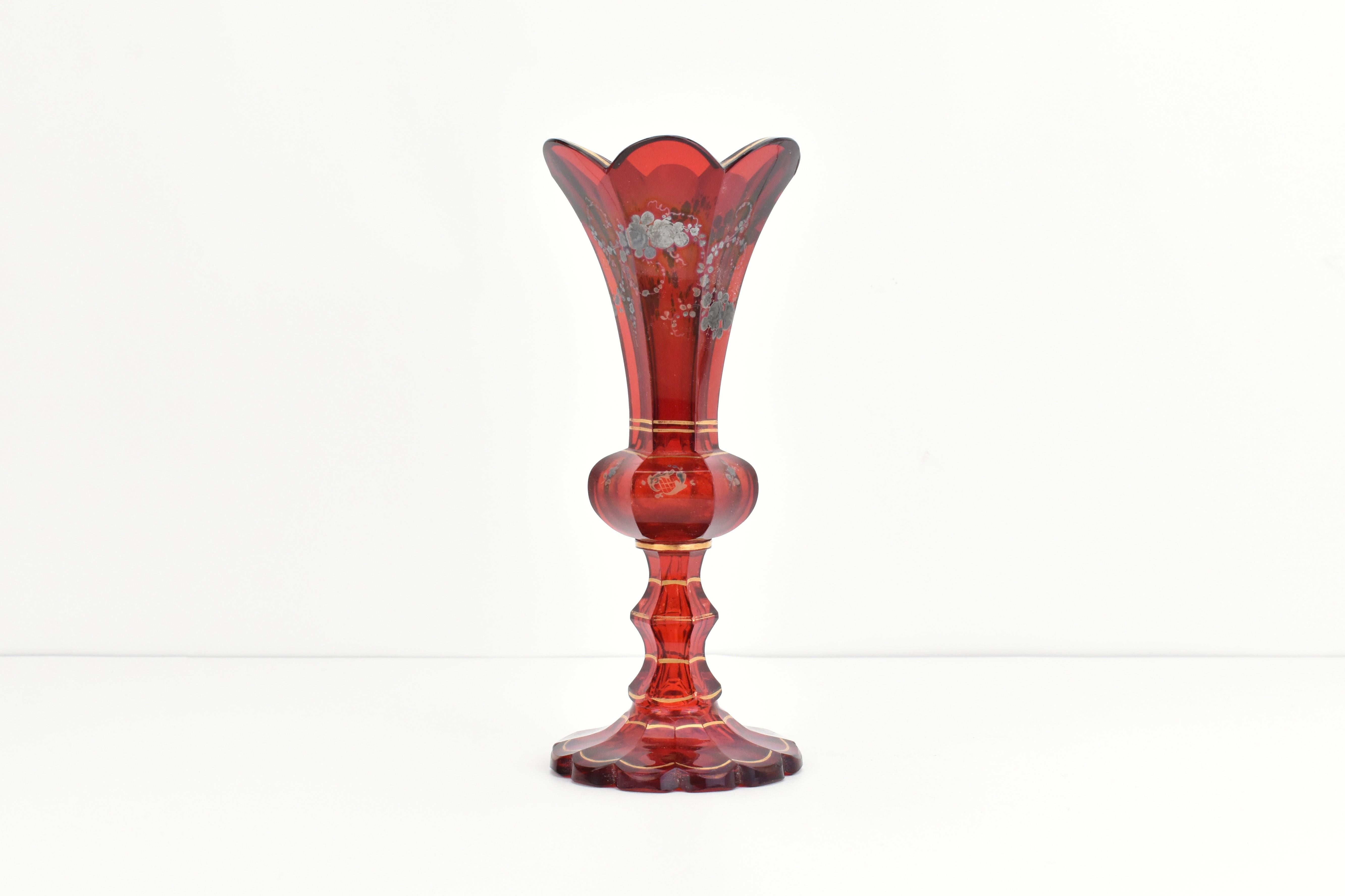 An elegant ruby red Biedermeier crystal glass vase, crafted with precision in Bohemia, circa 1850.

The body is beautifully cut into 6 side panels, each showcasing the vibrant ruby glass, and is adorned with hand-painted silver enamel featuring