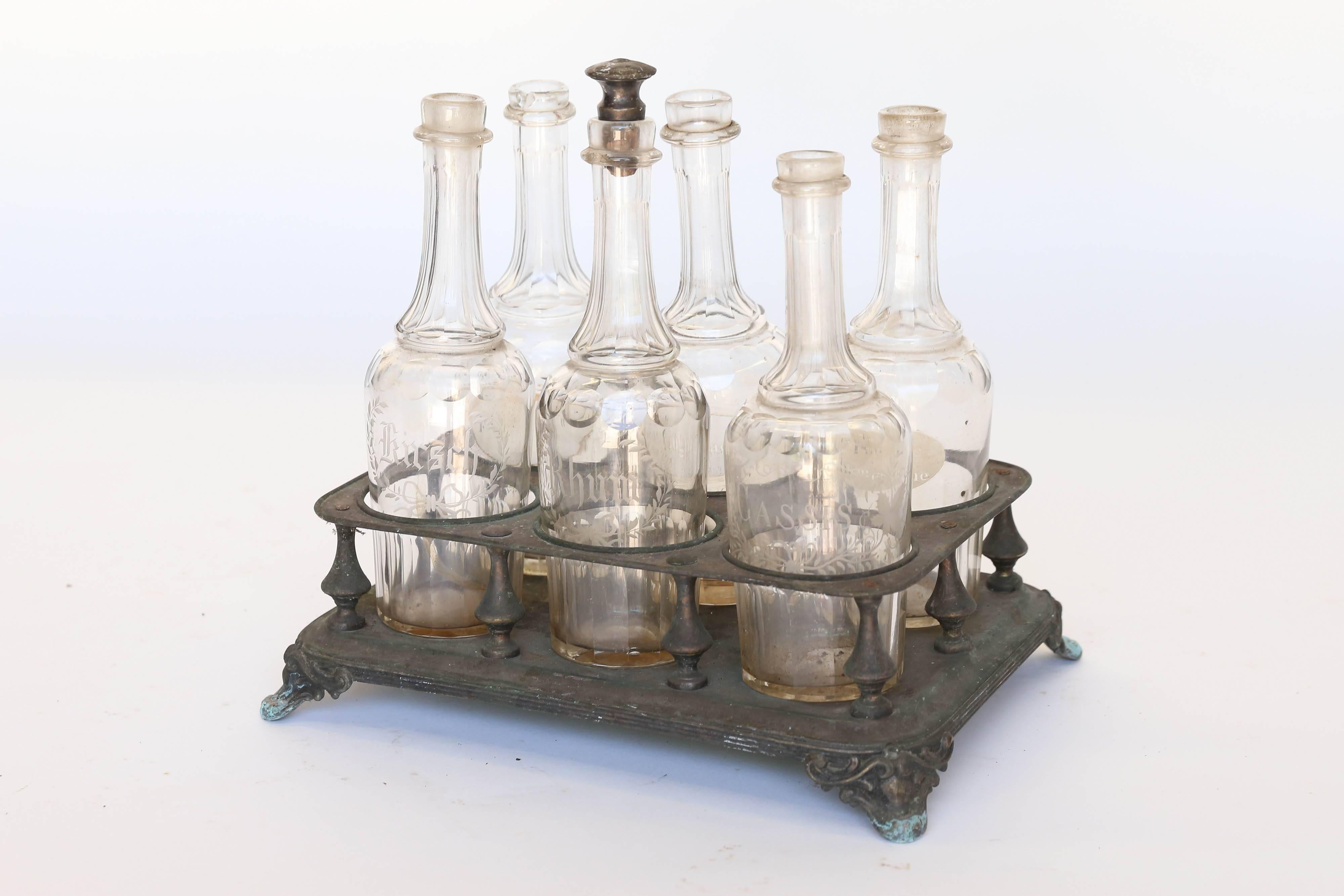 Found in France, a very old and lovely bottle holder with six etched bottles. Made of tin, the holder stands on four ornate feet and holds three liquor bottles elaborately etched 'Kirsch', 'Rhom', and 'Cassis'; the three remaining bottles are less