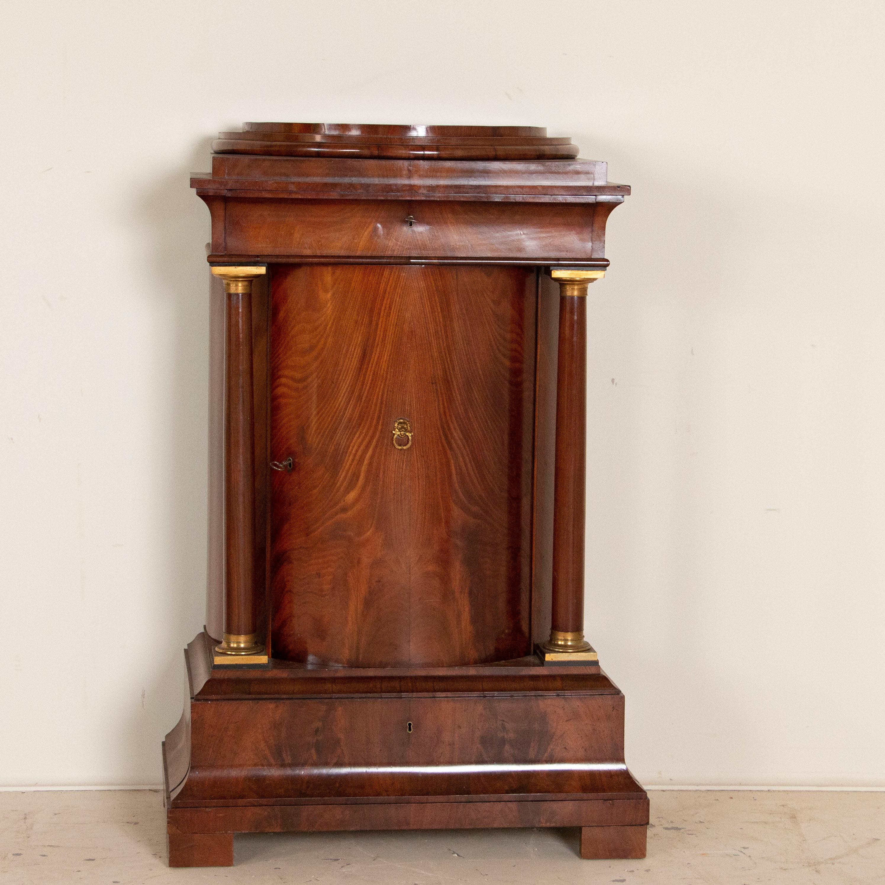 Stately and elegant, this mahogany cabinet reveals its Biedermeier style in the Classic columns, bronze accents and bowed/curved door, in addition to the striking finish of the rich mahogany. Designed as a pedestal cabinet by an exclusive craftsman,