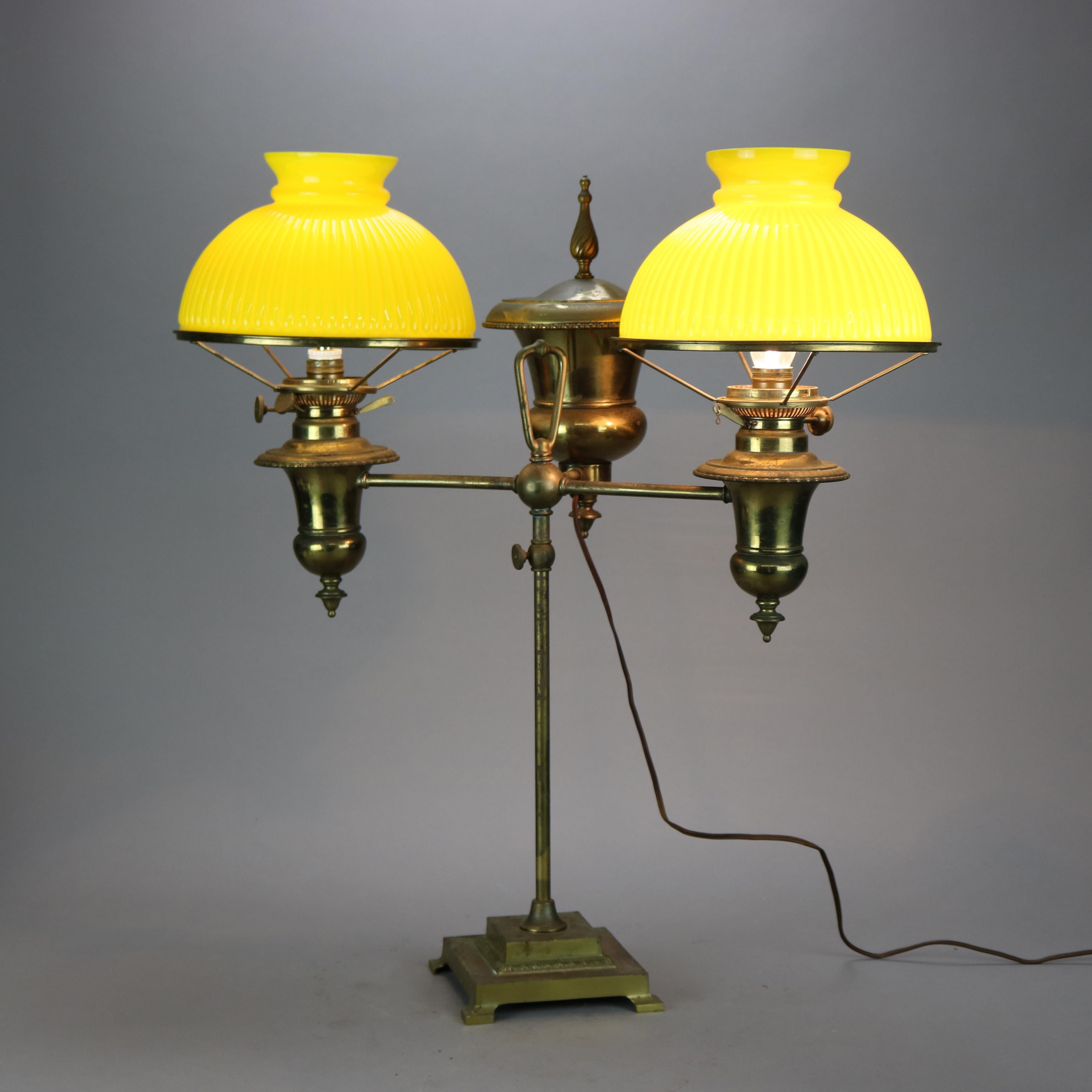 An antique double student lamp by Bradley and Hubbard offers brass frame with urn form fonts, one having stylized flame finial, on shaft with adjustable arms terminating on lights with ribbed yellow cased glass shades, electrified, maker mark as