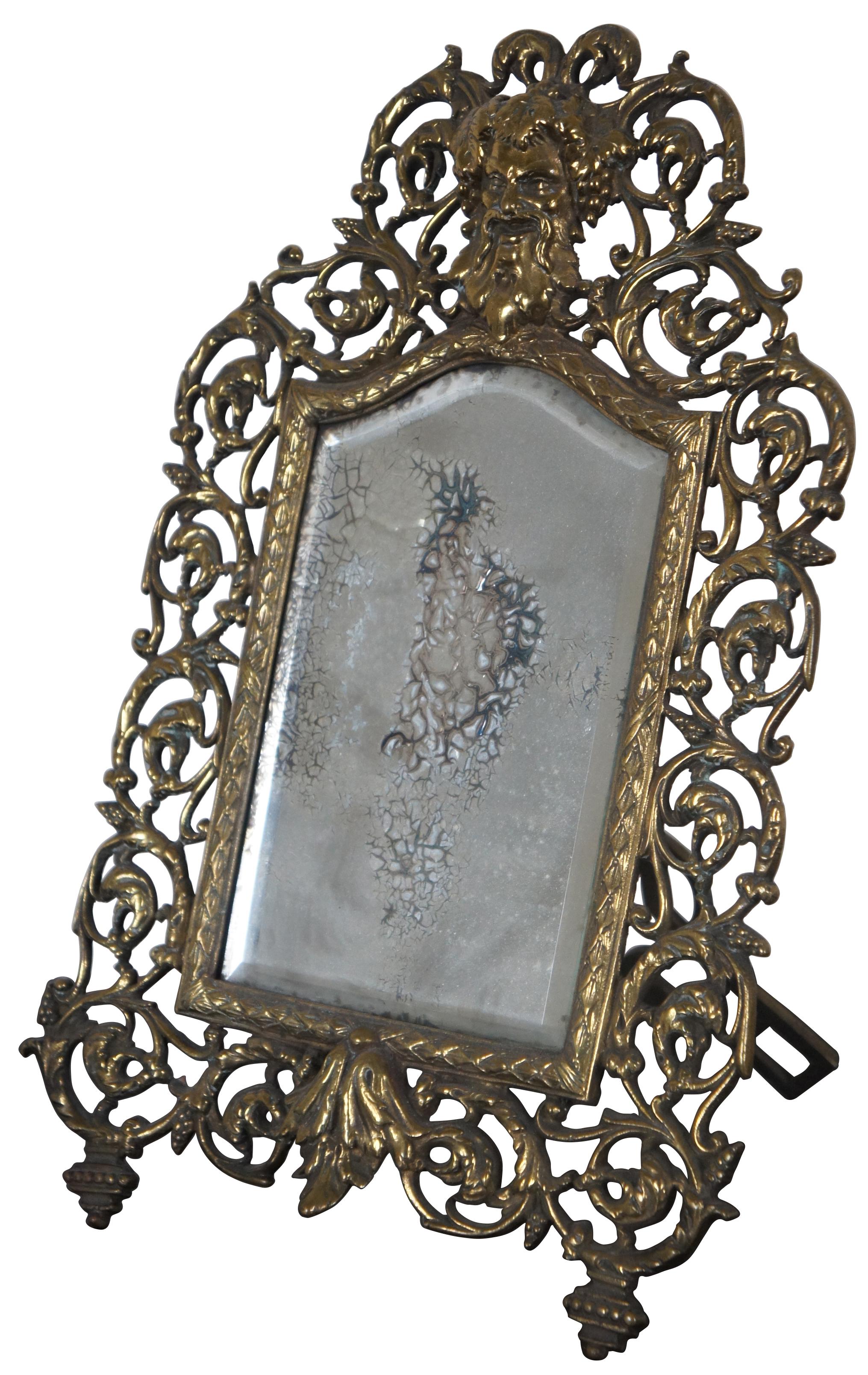 Antique gilded cast iron easel style tabletop mirror with an ornate frame of swirling leaves around the face of Bacchus or Dionysus. 

Measures: 10” x 8.5” x 14.125” / Mirror - 5” x 7.75” (width x depth x height).