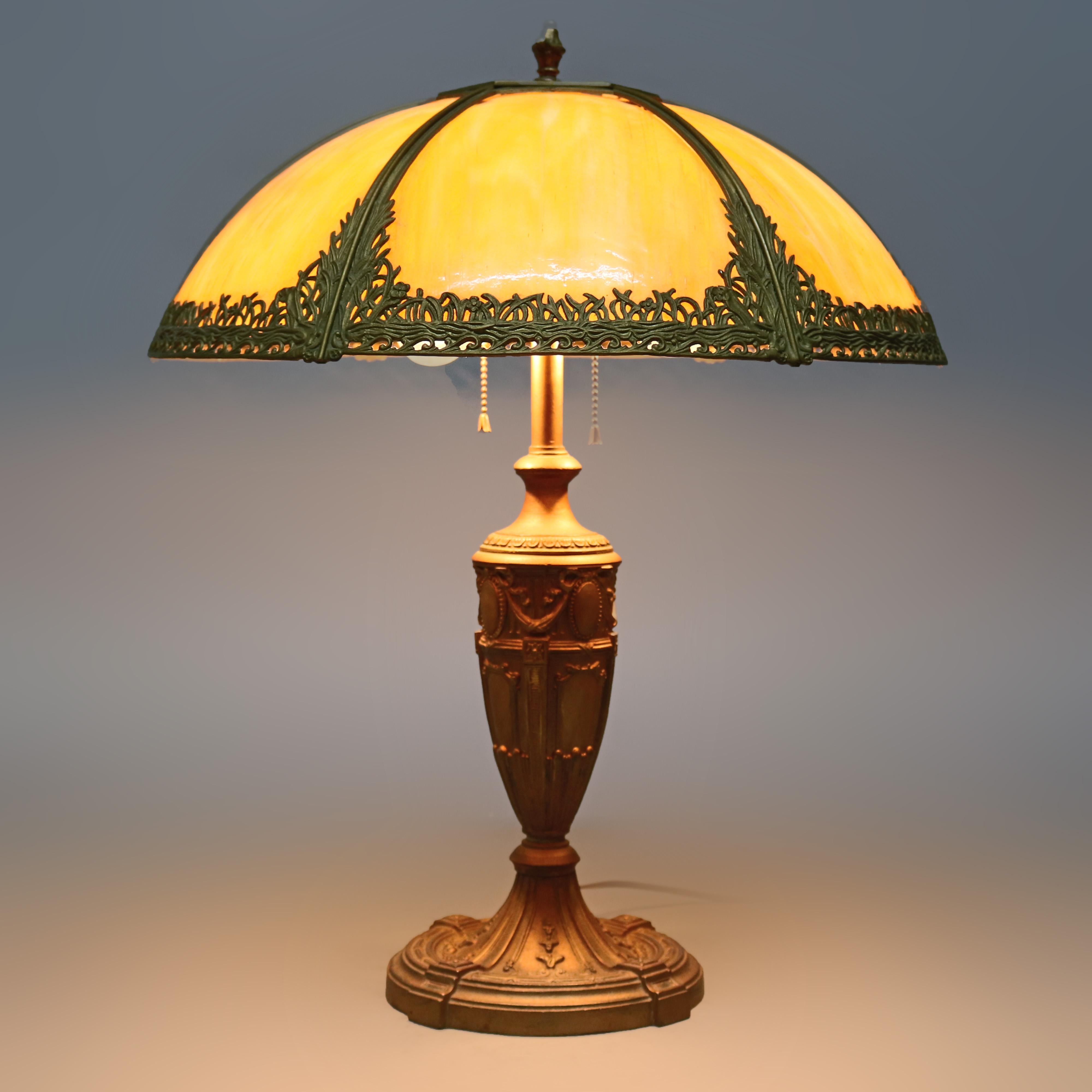 An antique Arts & Crafts table lamp in the manner of Bradley & Hubbard offers cast and gilt foliate filigree frame housing curved slag glass panels surmounting urn form double socket base with embossed Classical elements, circa 1920.

Measures: