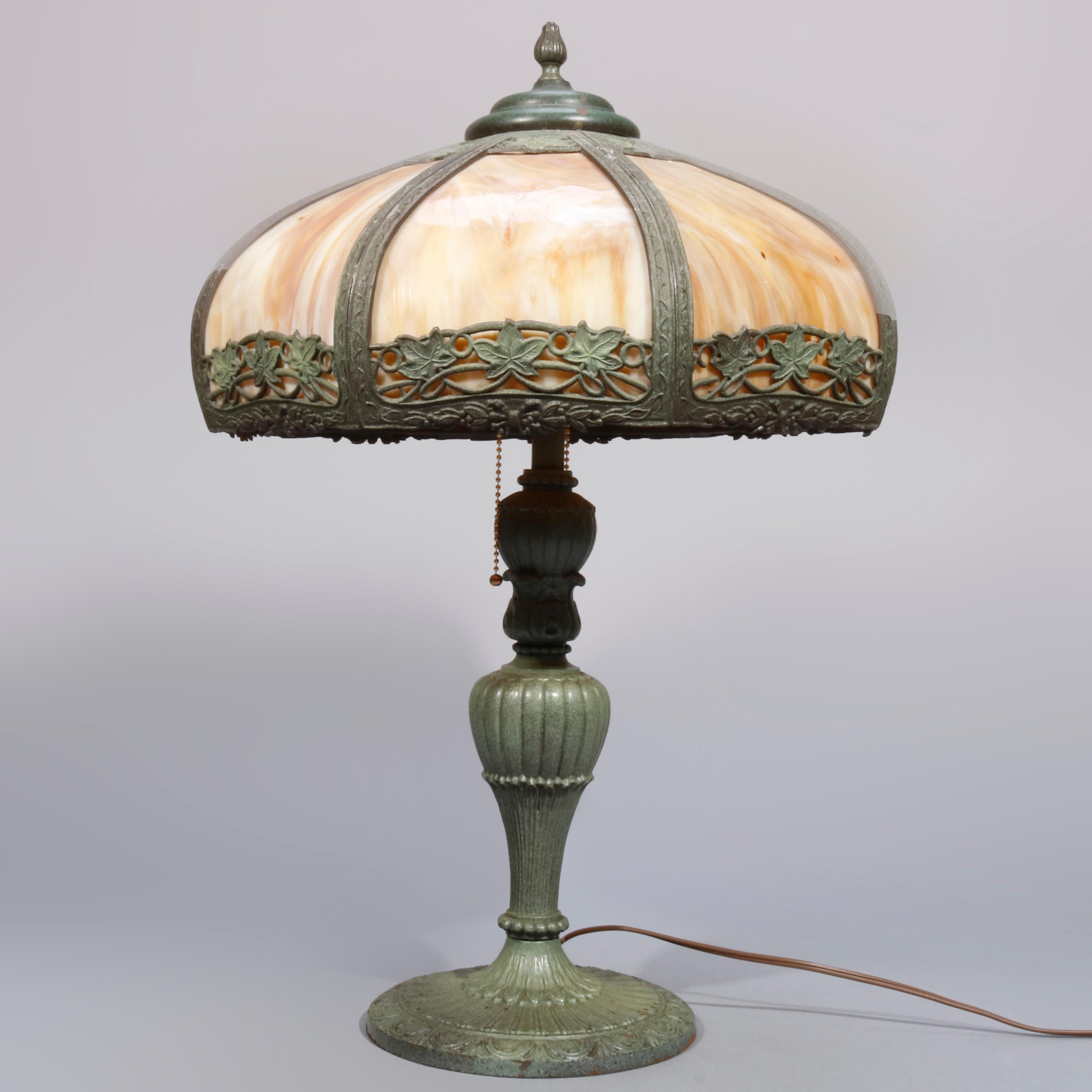 An antique table lamp in the manner of Bradley & Hubbard offers cast foliate filigree shade with verdigris finish and housing curved slag glass panels surmounting double socket cast urn form base, circa 1920

Measures: 23.25