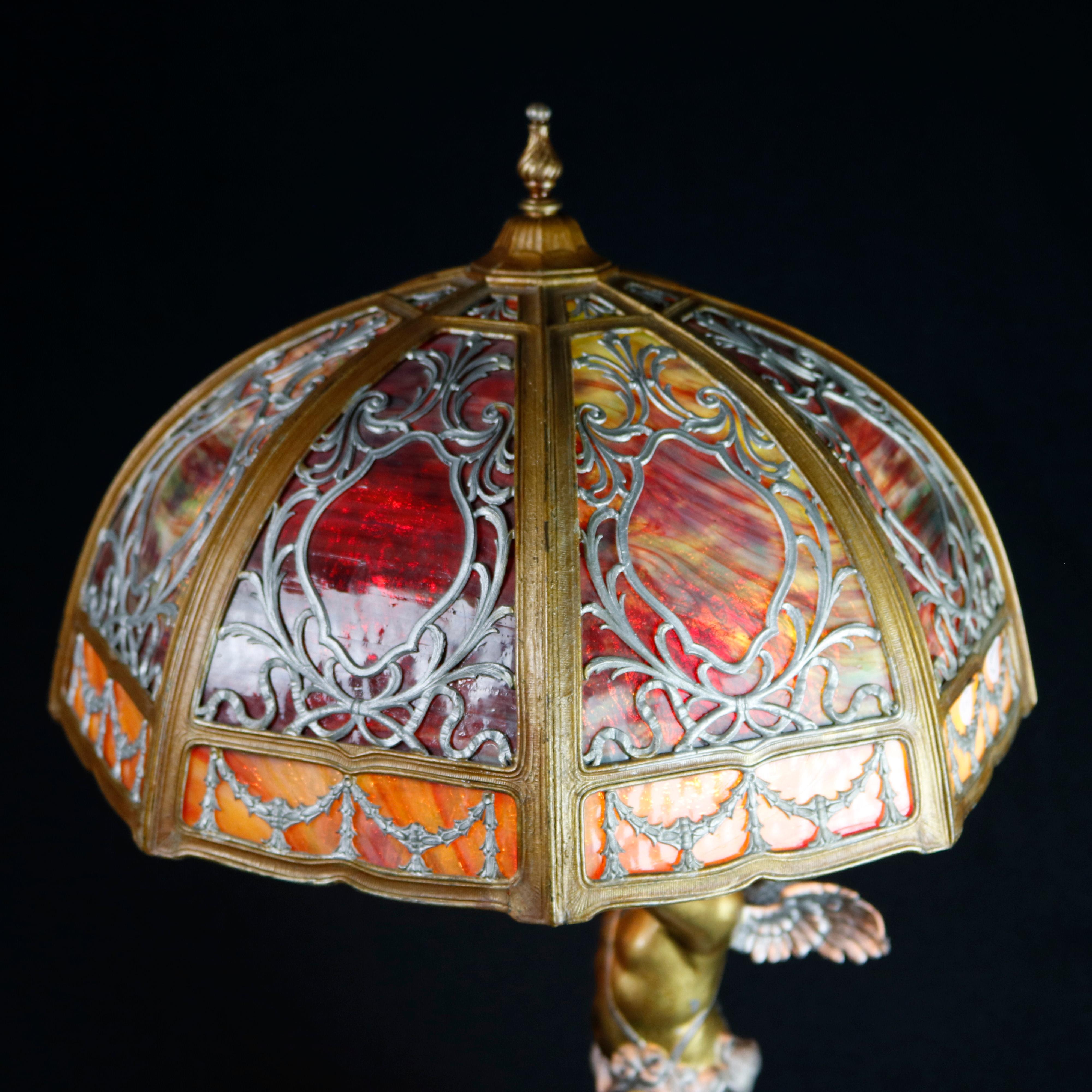 An antique figural Bradley & Hubbard School table lamp offers dome form shade with foliate, scroll and garland filigree shade frame having bent slag glass panels surmounting cast base with winged cherub on raised on embossed and footed gilt base