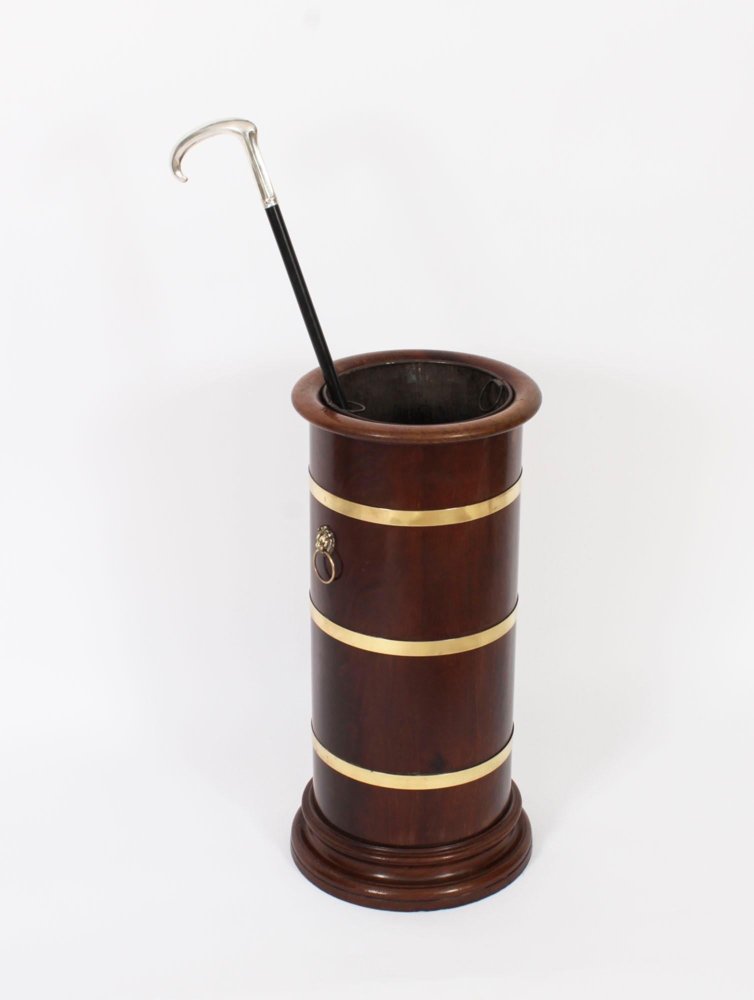 This is an elegant antique Victorian brass bound mahogany walking stick stand, circa 1870 in date. 

The stand is of cylindrical shape, features three brass bands, a pair of ormolu lion mask handles and has its original zinc lining.

This stand has