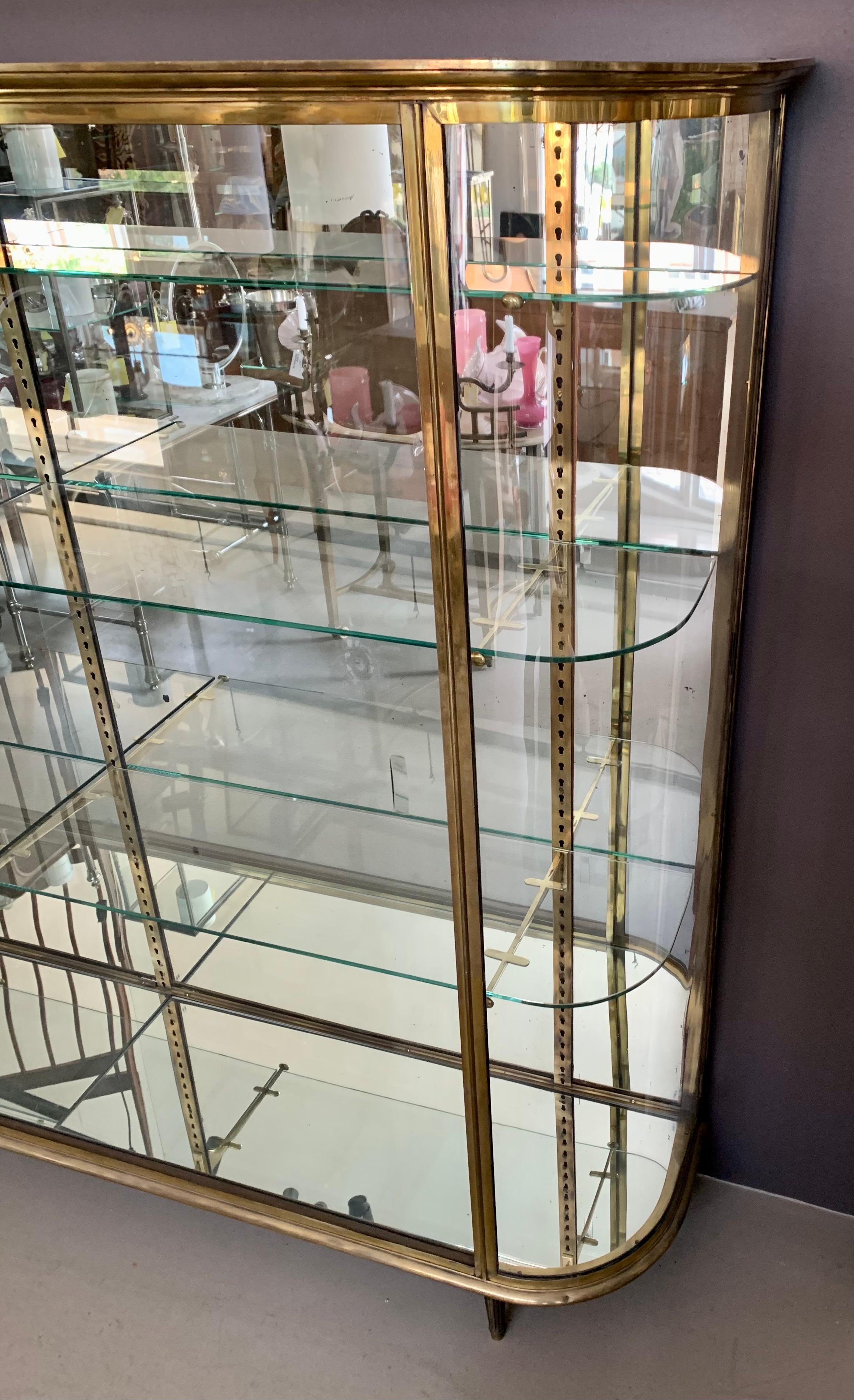 Excepcionalmente grande e impresionante vitrina francesa de latón antiguo con estantes de cristal, elegantes esquinas redondeadas y dos puertas correderas de cristal. El mueble, que sí tiene algo de peso, se levanta con gracia del suelo sobre patas