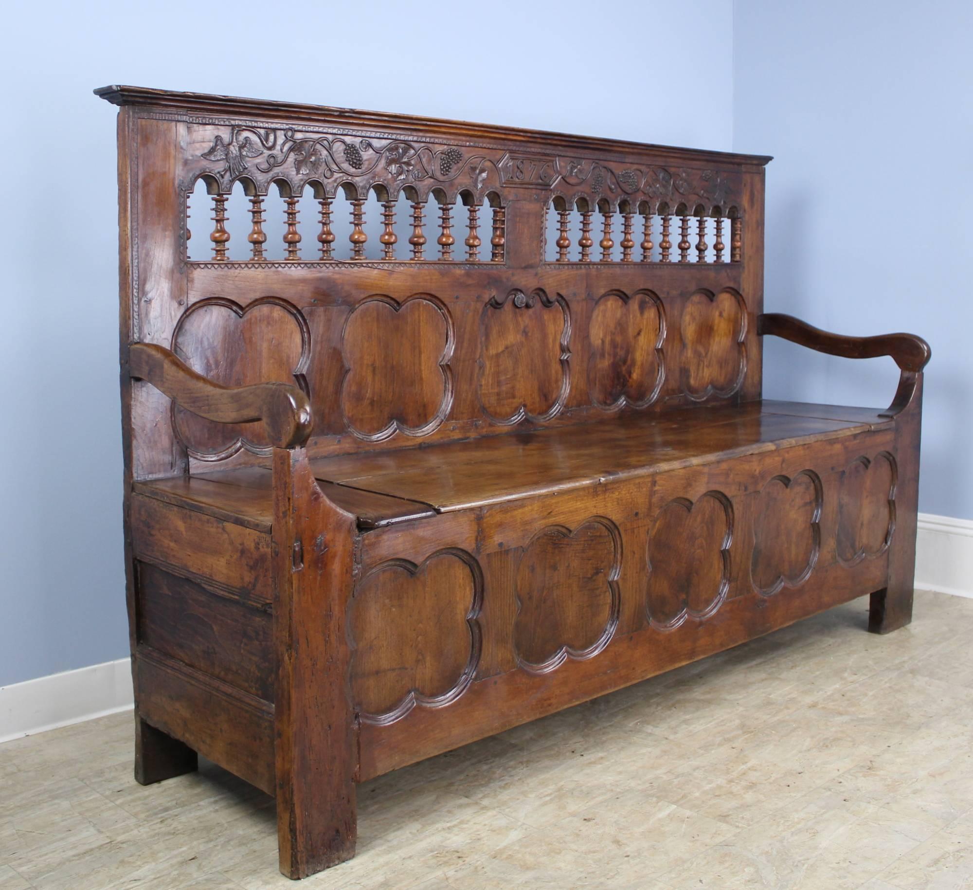 A fabulous ornately carved walnut bench from Coastal France. Charming spindles under a panel of hand carved birds, leaves and grapes, with the date front and centre. The seat lifts up (not hinged) to reveal a large storage area. The repeating clover
