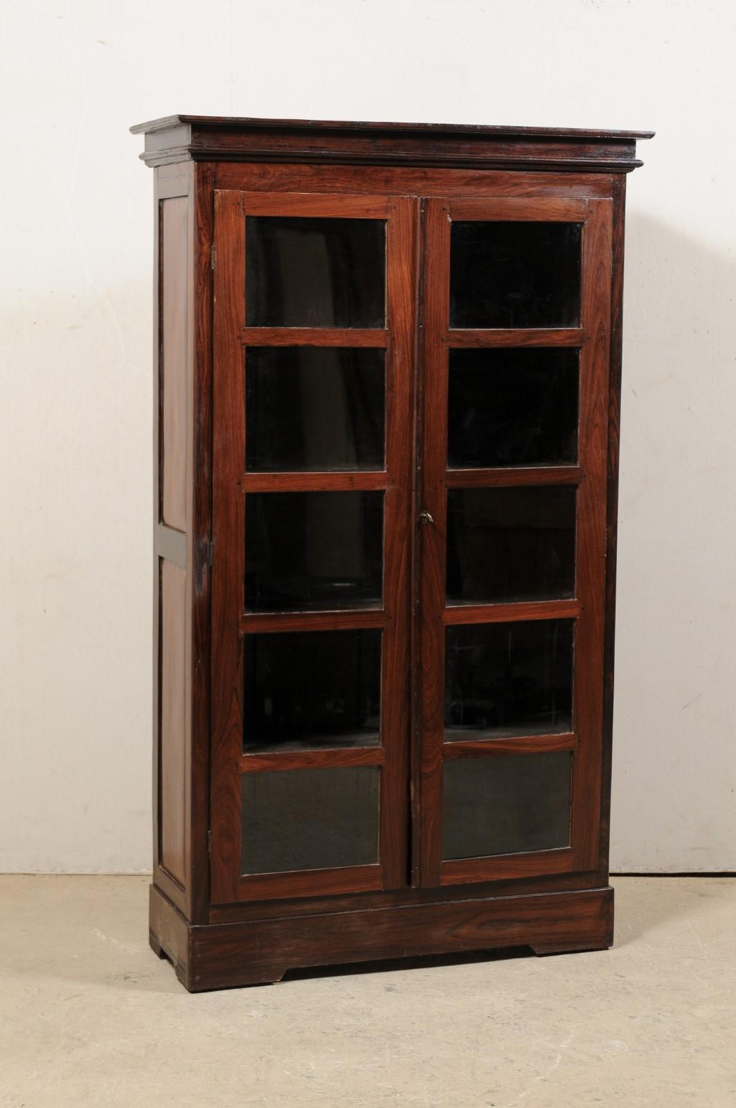 A tall British Colonial rosewood storage cabinet with glass doors from the early 20th century. This antique display cabinet features a molded cornice atop a tall case with pair of five-panel front doors, set with glass. The skirt and sides of the