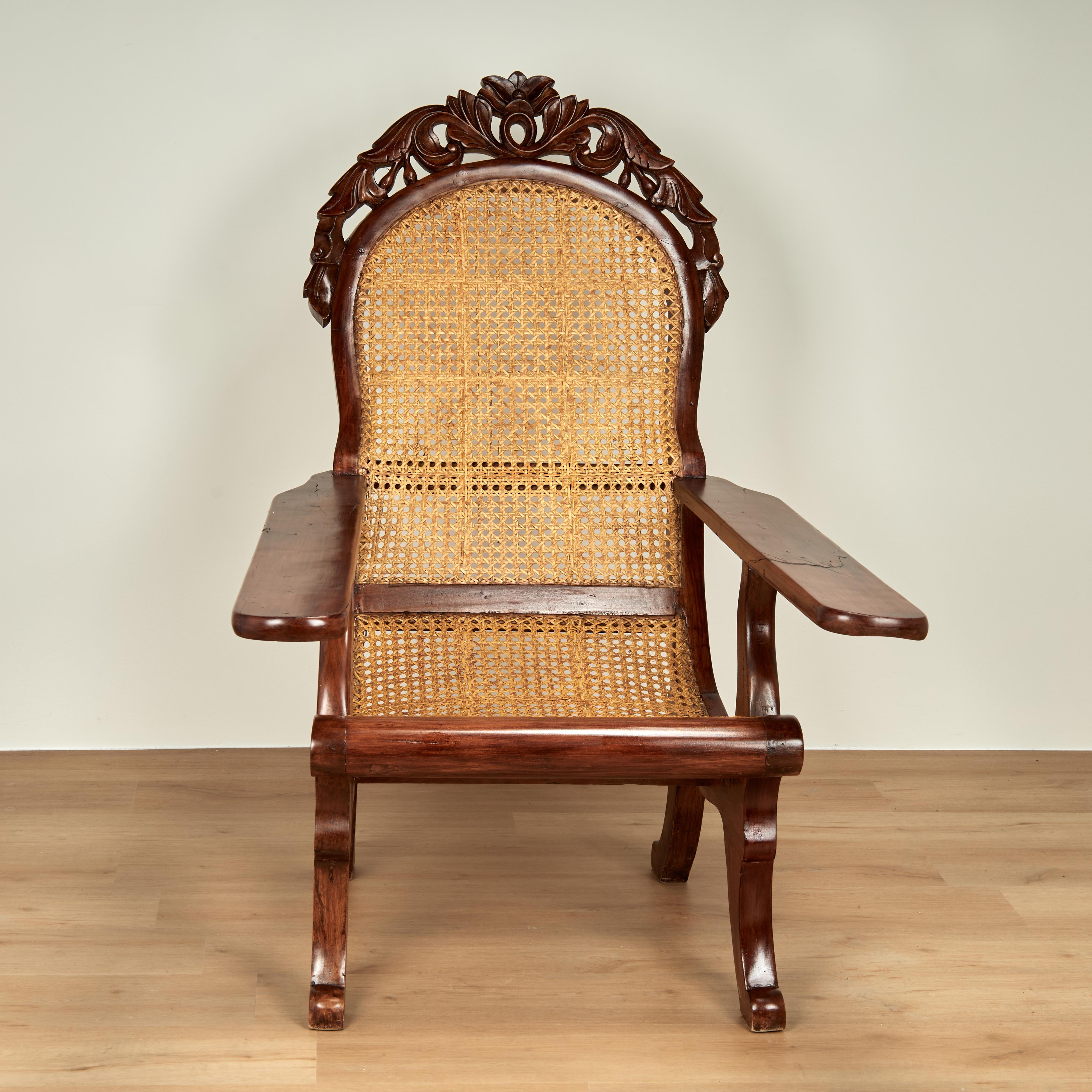 Transporting 19th century British colonial plantation chair handcrafted in mahogany with elaborate floral carvings on the crest, caned back and seat, plank arms and scrolled legs.