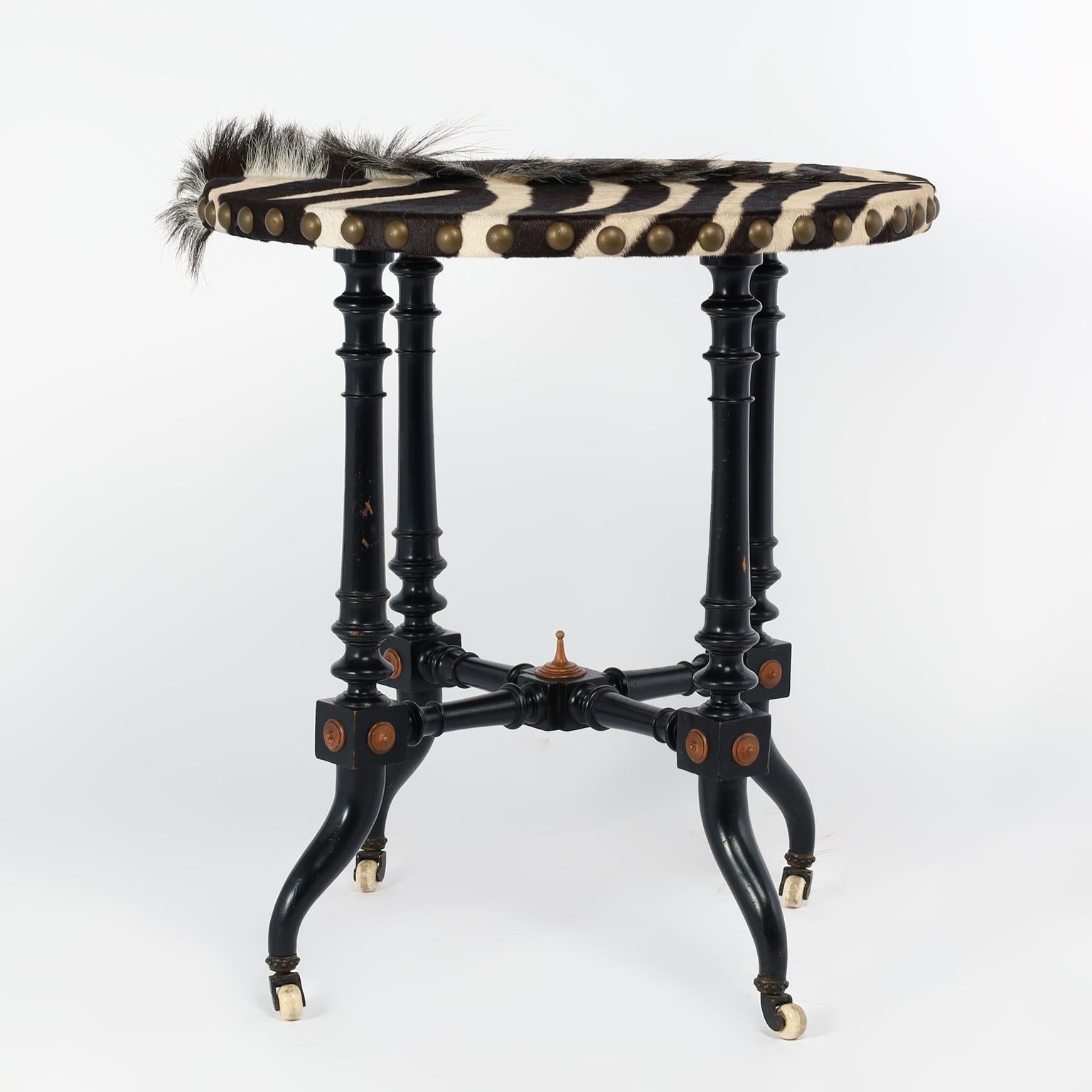 Rare and unusual 19th century English table or stand with a round top covered in zebra with brass tacks over an aesthetics movement base having four turned legs connected by cross stretchers on curved legs with casters on porcelain wheels.