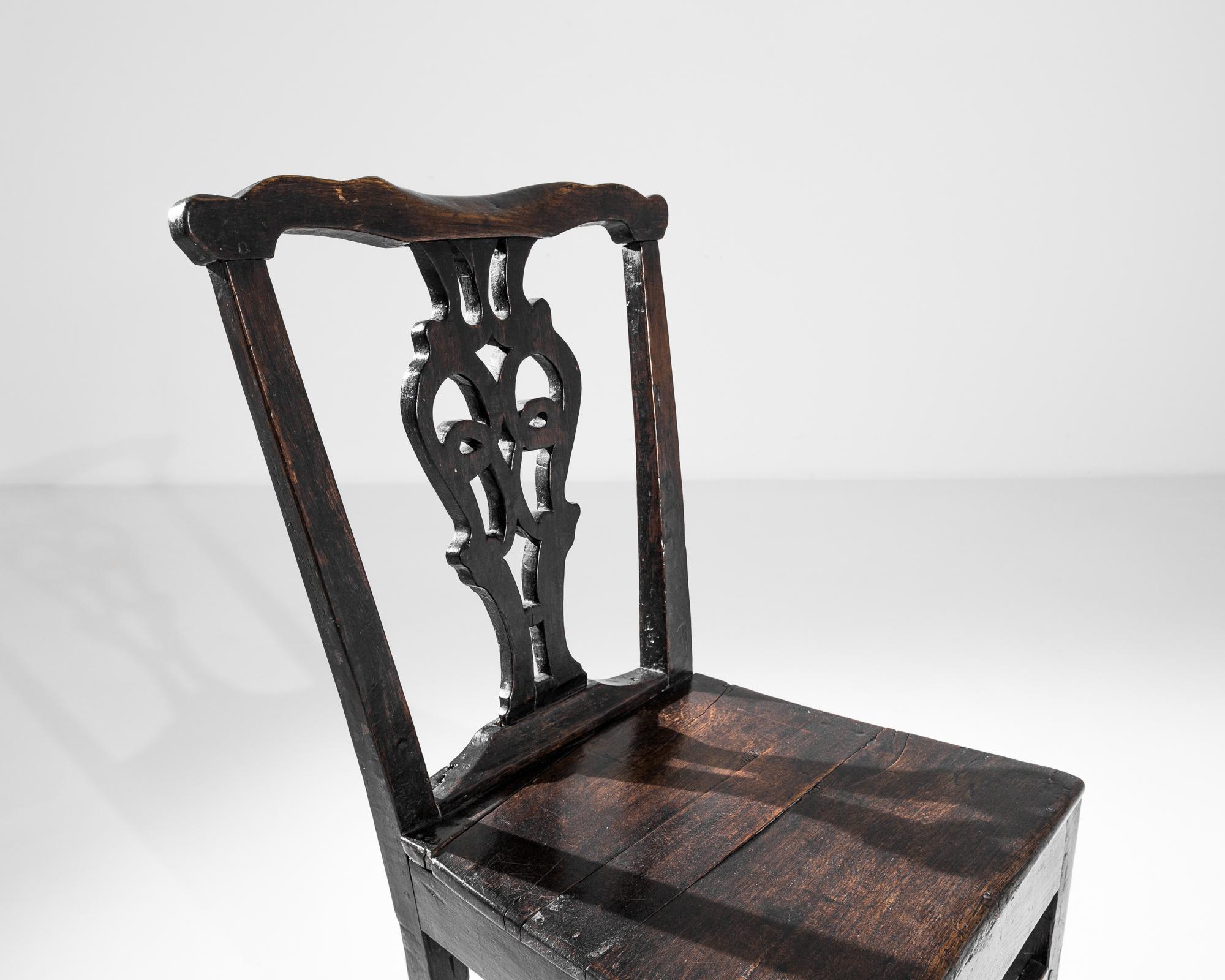 An ornate back splat and expressive patina give this oak chair a poetic character. Made in the UK circa 1800, the wood has retained its original finish: a deep ebony hue, brindled with amber inflections. The right-angled, upright seat finds an