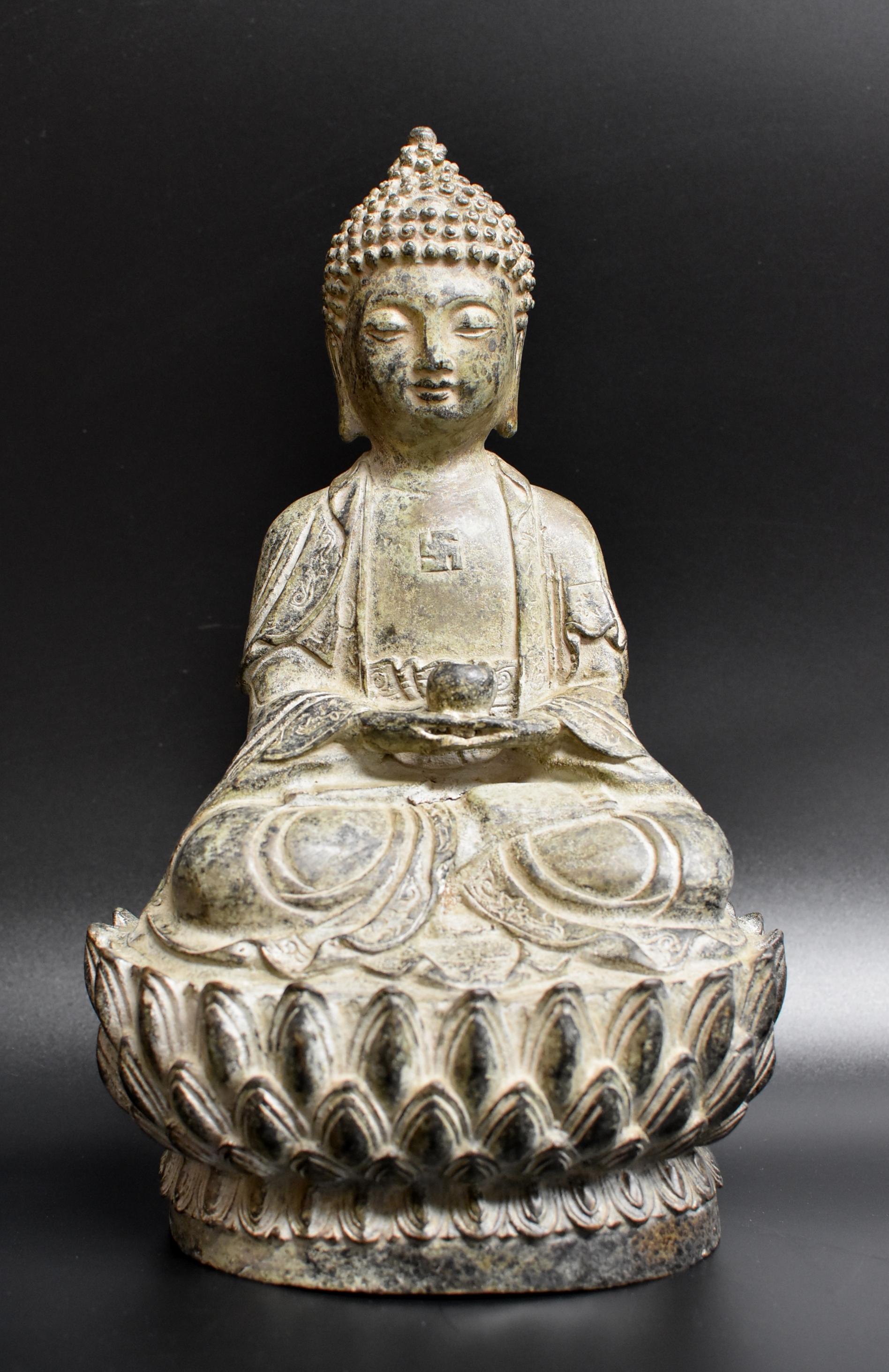 A bronze statue of Buddha seated on double lotus base. Small, youthful face with arched eyebrows and downcast eyes casting a serene aura, framed by long-lobed ears and an ushnisha symbolizing wisdom and attainment of enlightenment. Slightly opened