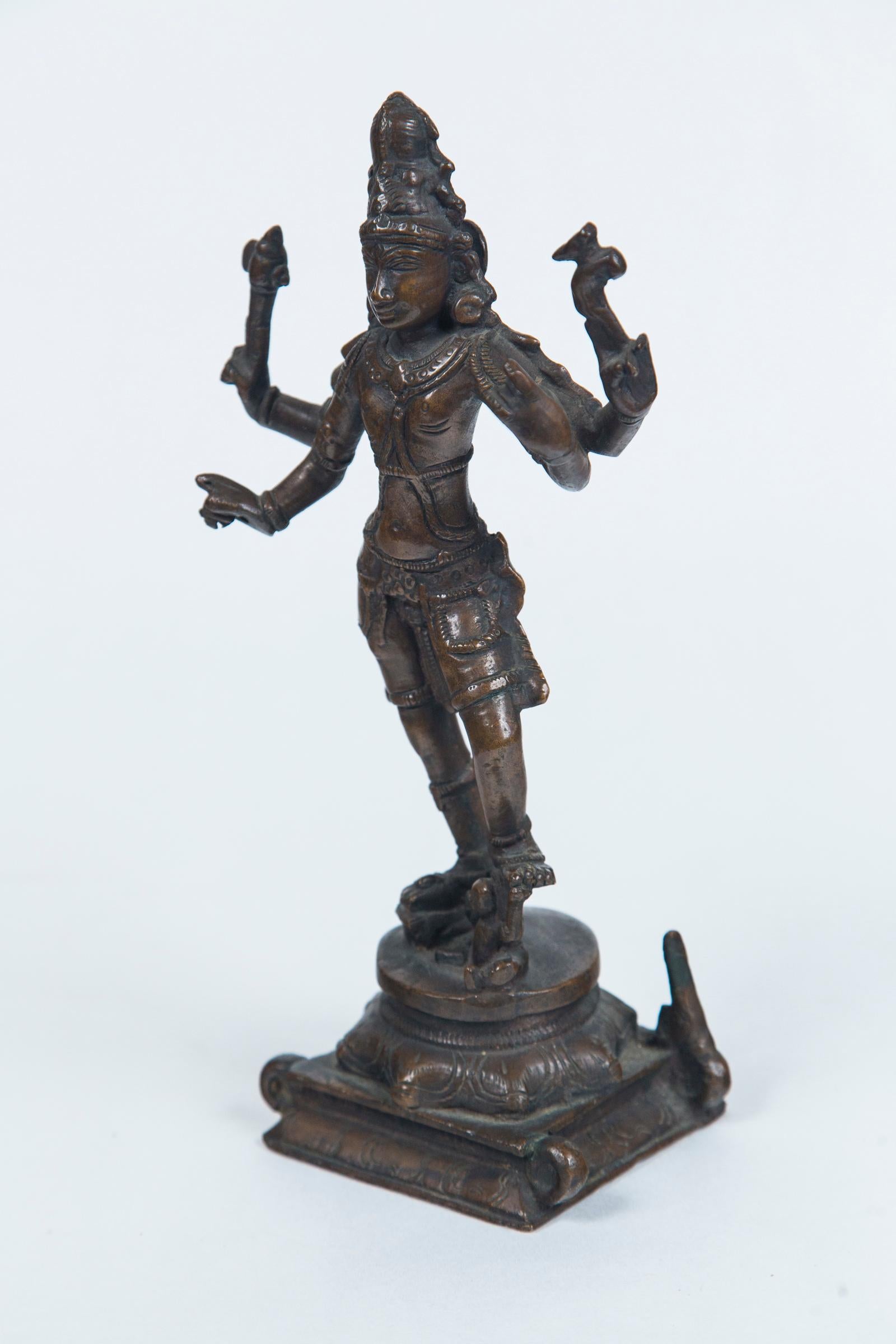 The 4 armed bronze Shiva holding symbols of his powers. His left foot rests on a demon. He wears an ornate headress. Dark natural patina. Raised on a square base.