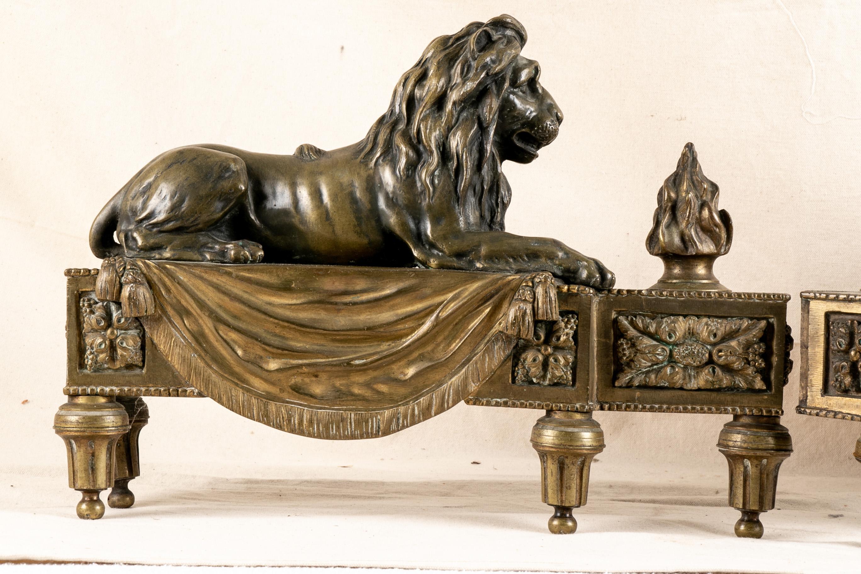 Antique bronze lion form chenets, recumbent lions on drapery covered plinths with tassels. Relief panels with leaf and berry motifs and three-dimensional flame motifs on the front ends. Raised on fluted peg form legs. Hollow backs. 

Condition: