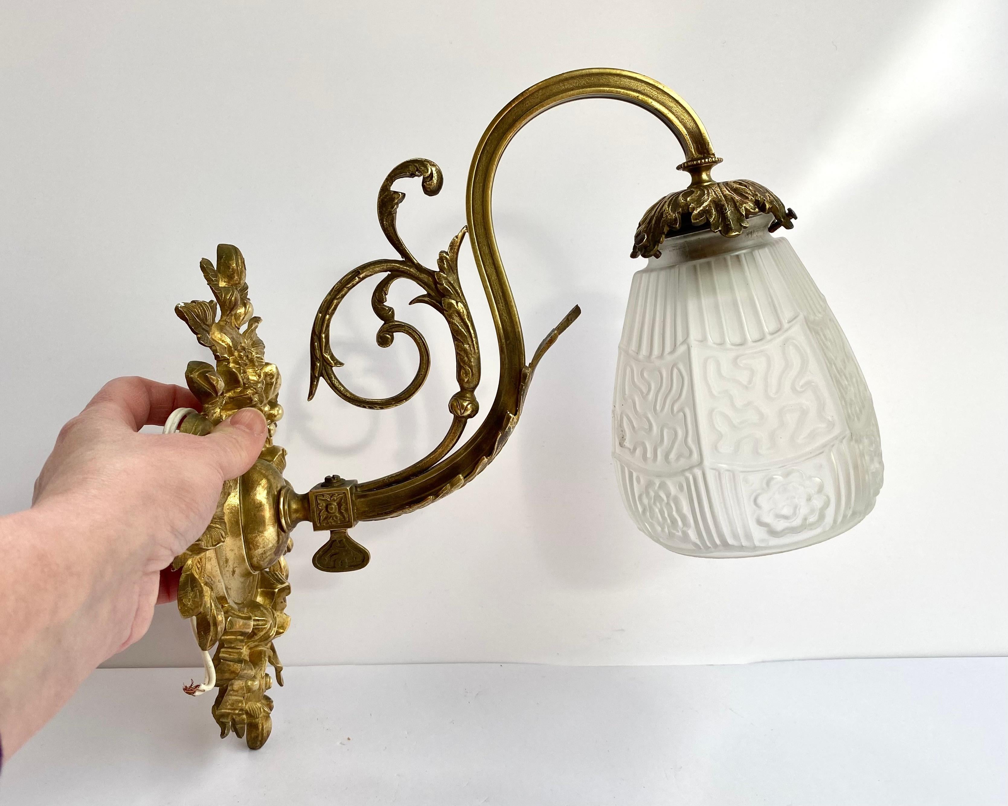 Elegante und stilisierte Jugendstil-Wandleuchte aus Bronze mit einem Lampenschirm aus weißem Glas in Form einer Blume.

Diese antike Wandlampe aus den 1930er Jahren ist mit aufwendigen, für die viktorianische Ära typischen Bronzegüssen