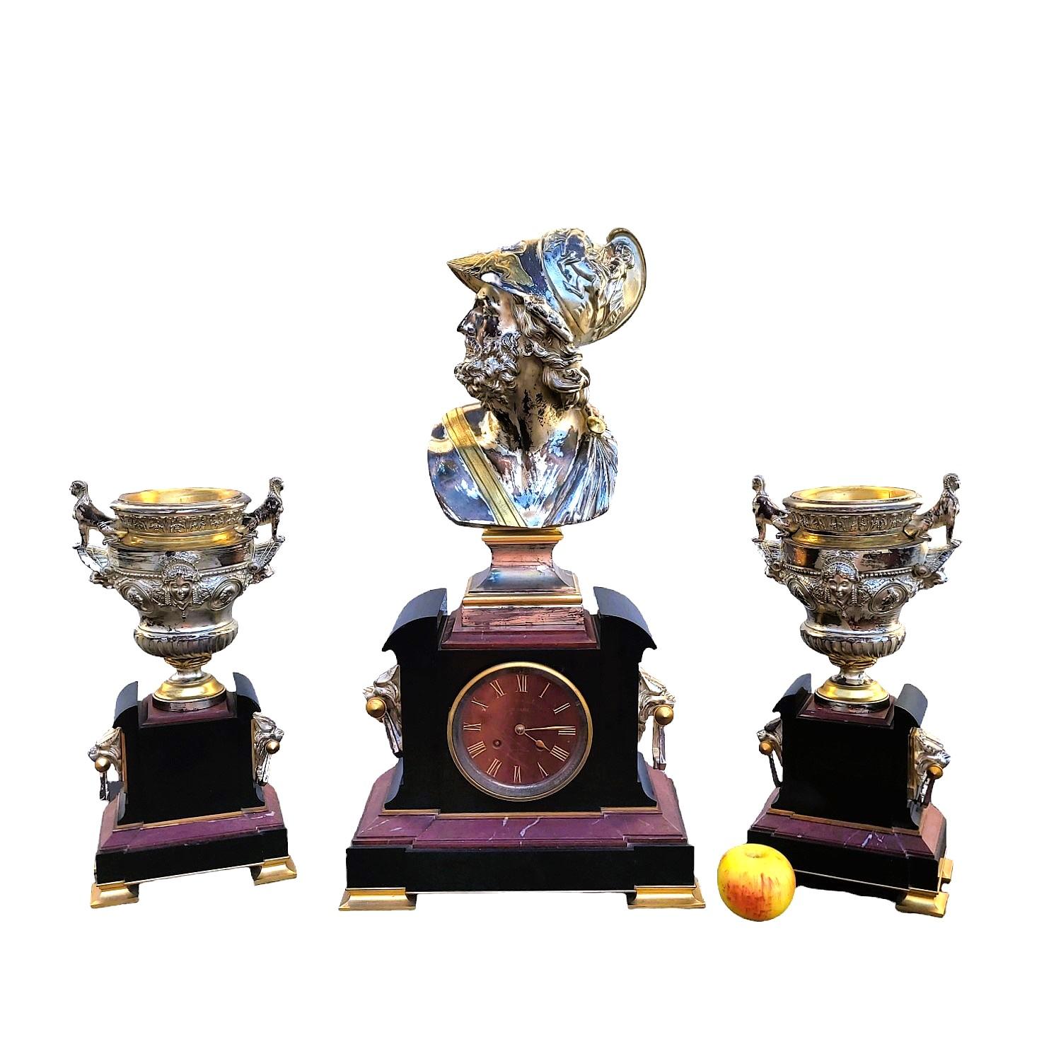 Conjunto de reloj compuesto por 3 piezas, un reloj y 2 urnas, de bronce patinado en oro y plata, sobre una base de mármol negro y rojo.

Inspirado en el neogriego, este conjunto presenta numerosas referencias a la mitología y al mundo griego. El