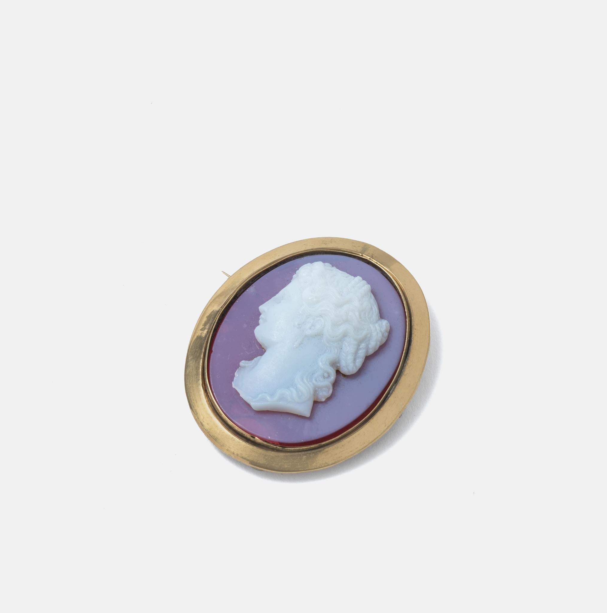 A bright and happy brooch which also can be used as a pendant with a chain. The oval central part is made of glass and has a beautiful woman portrayed in profile, white on a purple background. The frame around is made of gilt brass. Brooches were