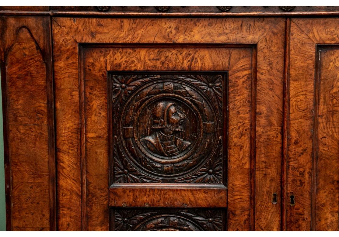 With a mirrored backsplash and a shelf and brass 3/4 gallery and supports. The top banded burled wood over a carved frieze in a Neo-Gothic style band. The double doors are carved with oak portrait medallions. Lacking a shelf and a key. The sides