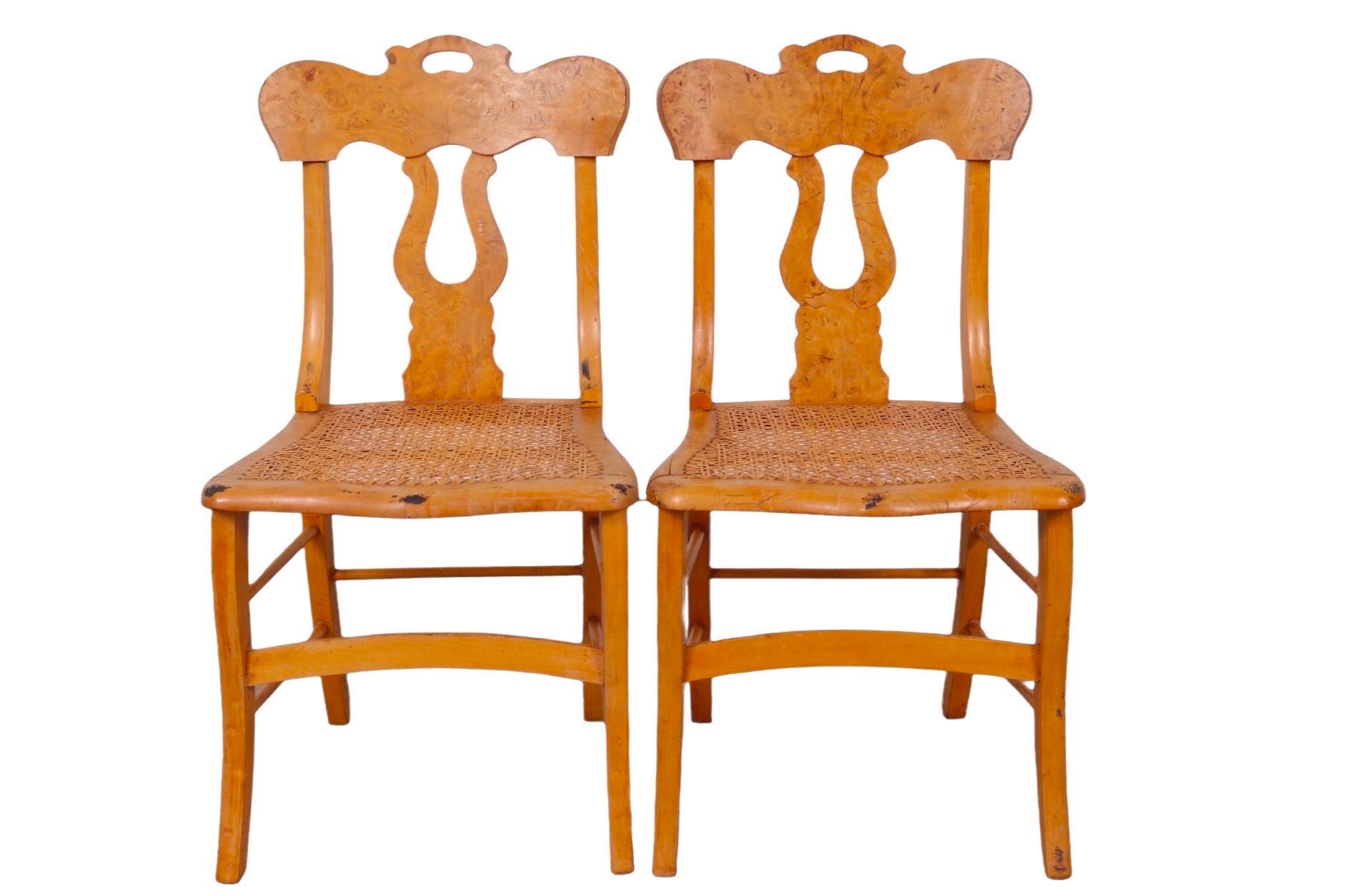 A pair of empire style side chairs dating from the late 1800’s. Seat backs are decorated with perched crest rails and lyre back splats covered in rich burlwood veneers. Hand caned seats are raised on saber legs, supported with box stretchers.