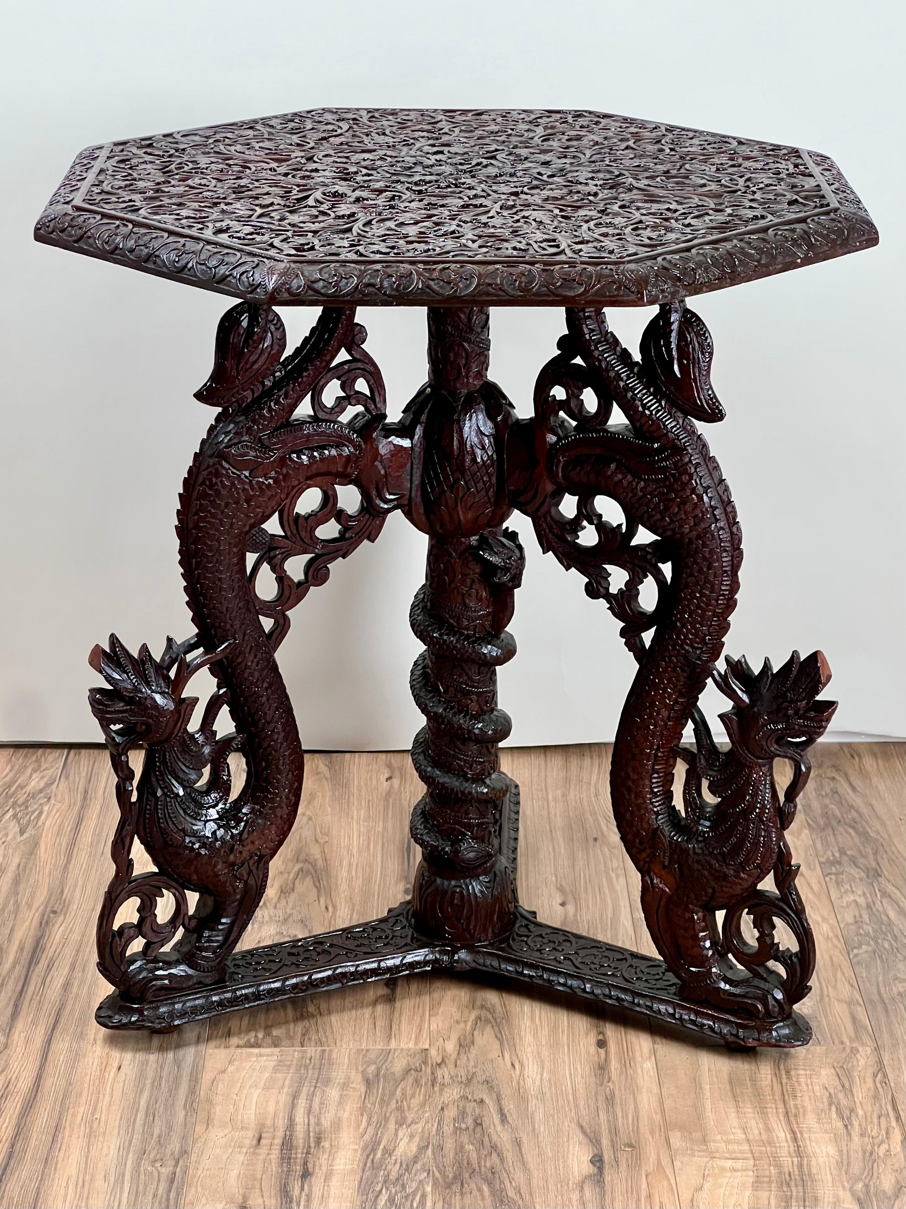 A masterwork of 19th-century Burmese craftsmanship, this intricately hand-carved table features an octagonal top supported by three dragons entwined around a central serpent column. The surface is richly ornamented with openwork floral and foliate