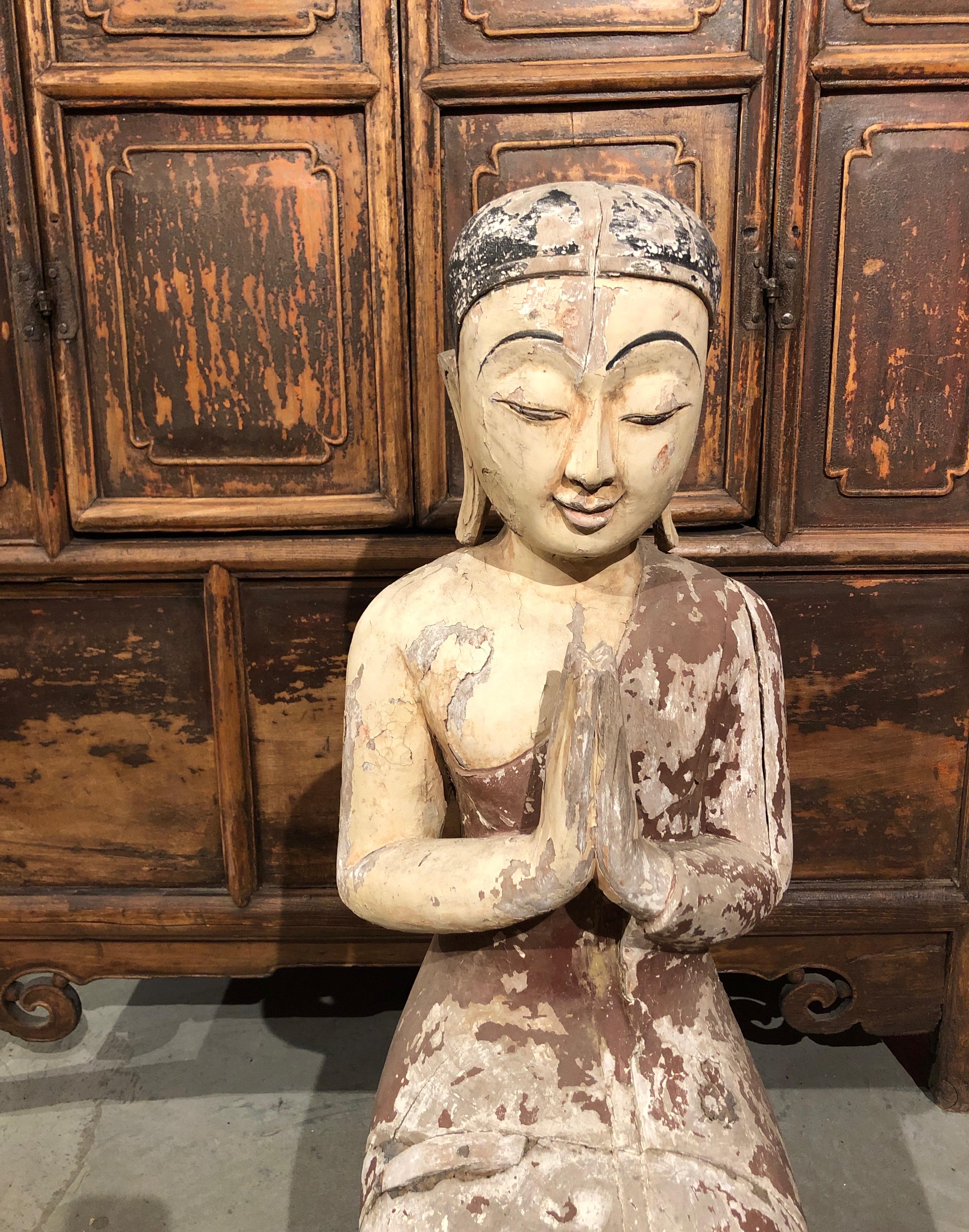 An antique kneeling monk on lotus leaf sitting in a praying position. From a Burmese Buddhist Temple. This well carved teak monk features attractive remnants of faded original paint and a striking aged patina from years in the sun. A truly evocative
