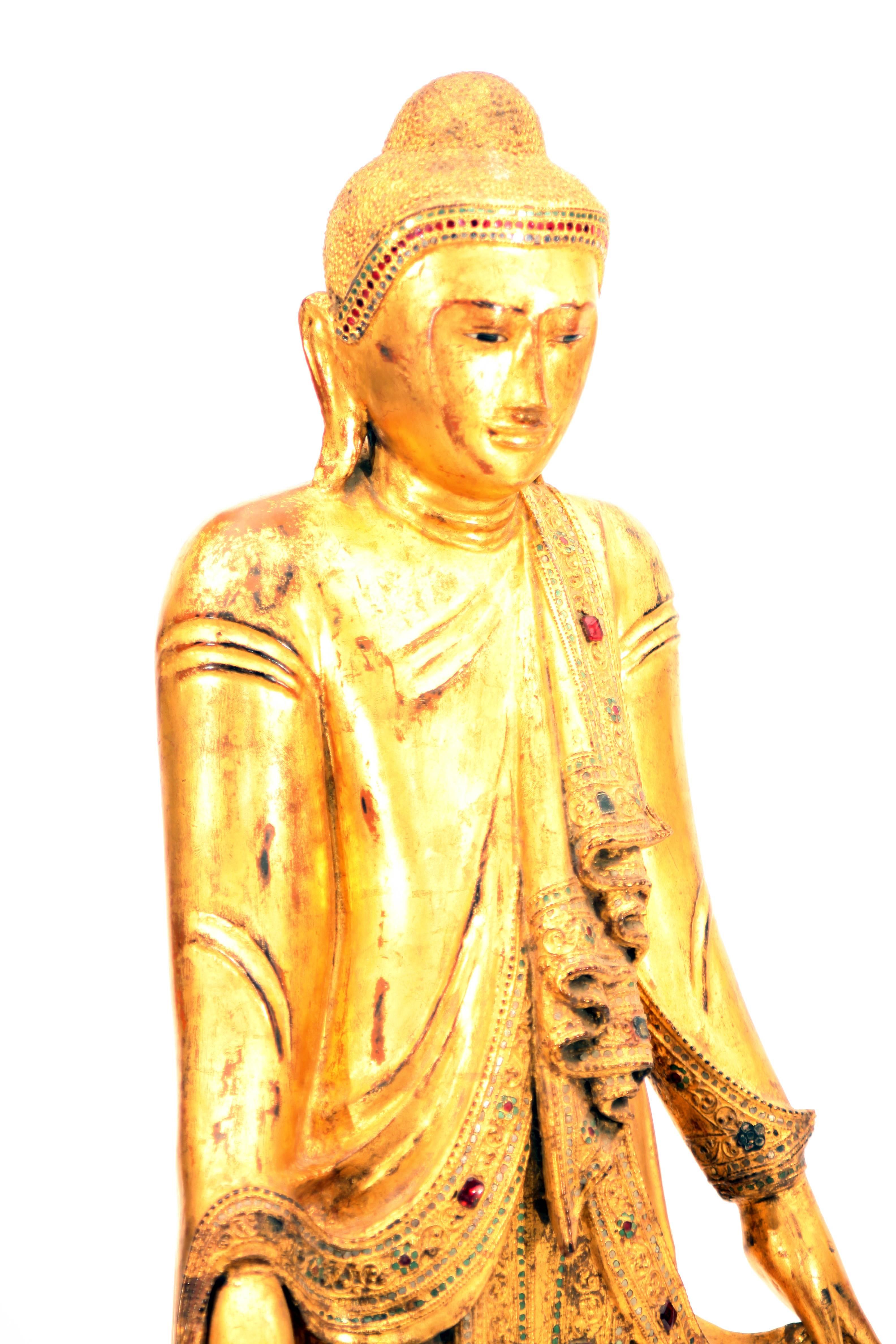 Standing on a lotus pedestal, with both hands clasping his robe, finely carved with flows and frills of robe and decorated with colored glass, the Buddha with serene expression, downcast eyes below arched eyebrows, aquiline nose, smiling lips,