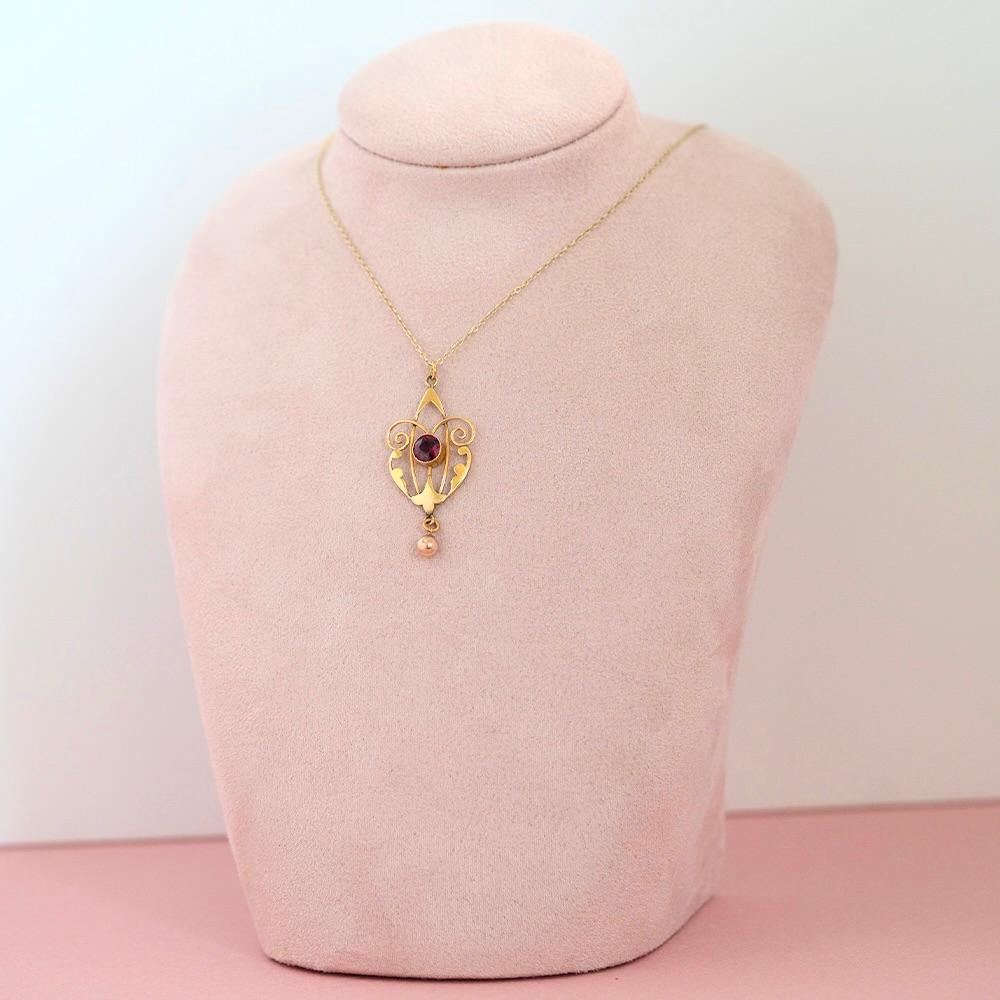 This elegant pendant captures the essence of Edwardian design, crafted in warm 9ct yellow gold with graceful openwork scrolls and soft symmetry. At its centre rests a richly hued round garnet, its deep wine-purple red tone glowing against the golden