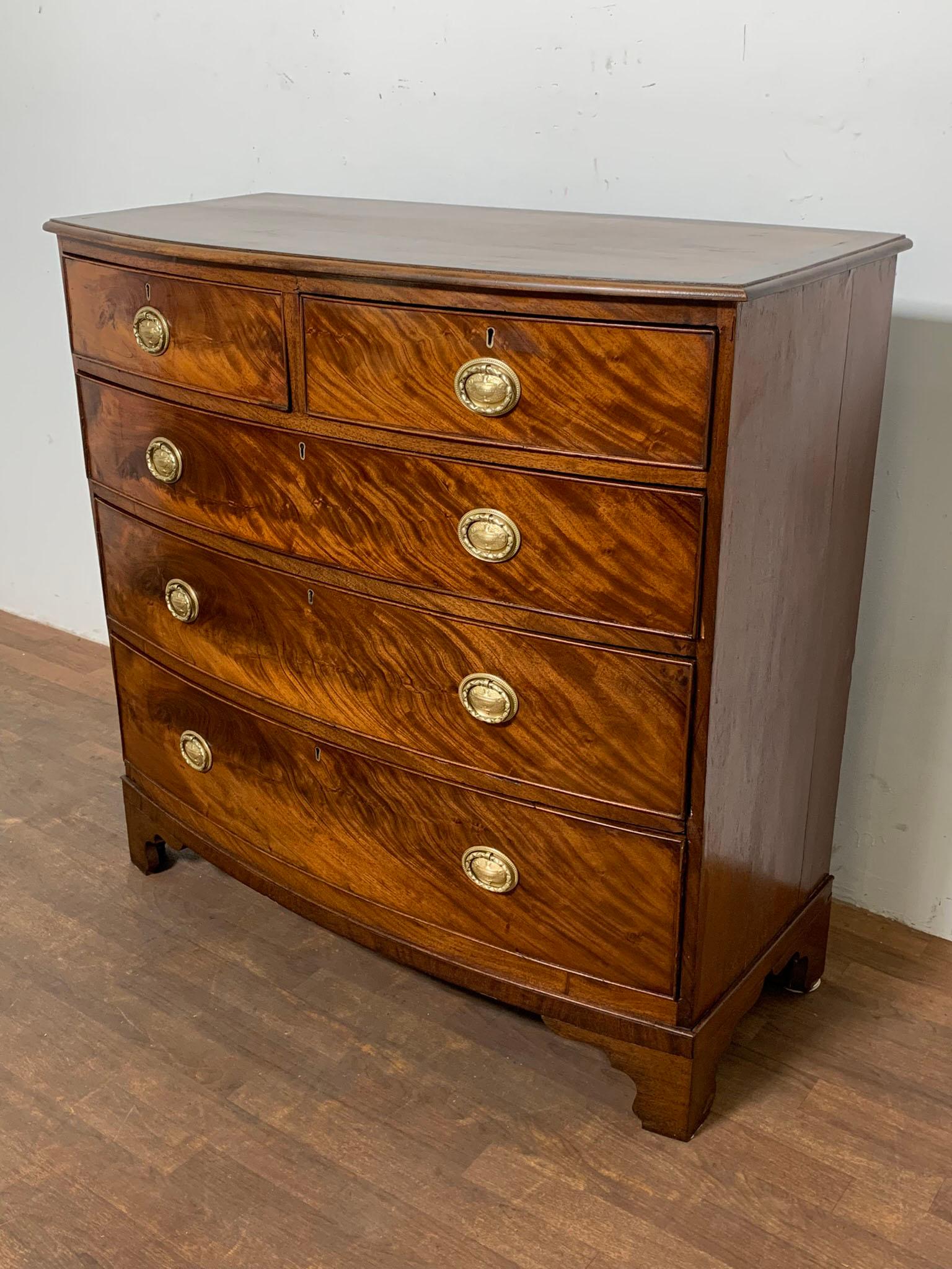 A George III era bow front chest of two over three graduated drawers in flame mahogany, made in England ca. 1800. Bears the stamp of the original early 19th century London distributor.