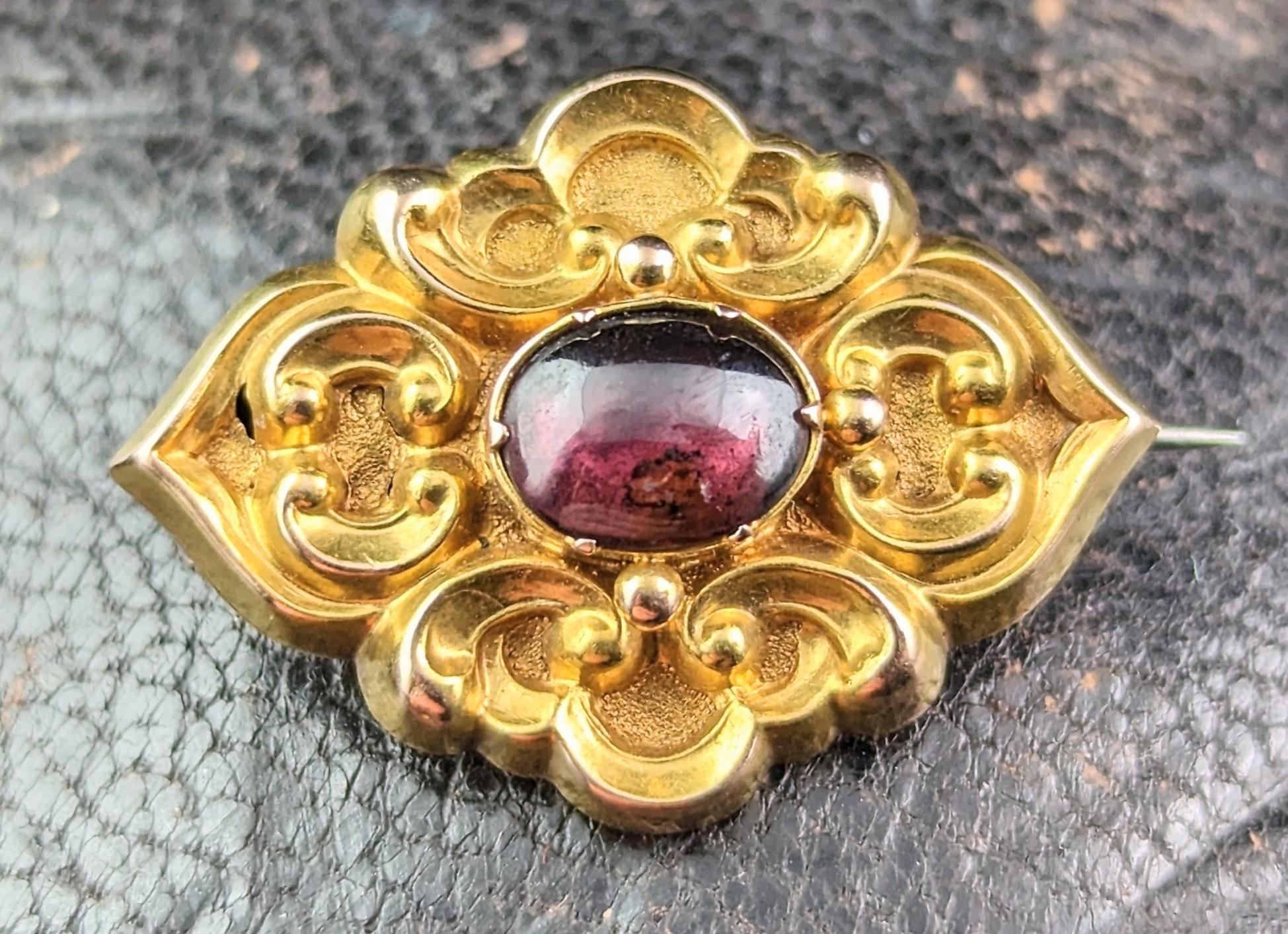 This antique Mid Victorian Garnet Cabochon mourning brooch is sumptuously beautiful.

Rich buttery 15kt bloomed yellow gold, the repousse scrollwork front housing a big juicy red garnet cabochon to the centre.

To the verso the brooch has an