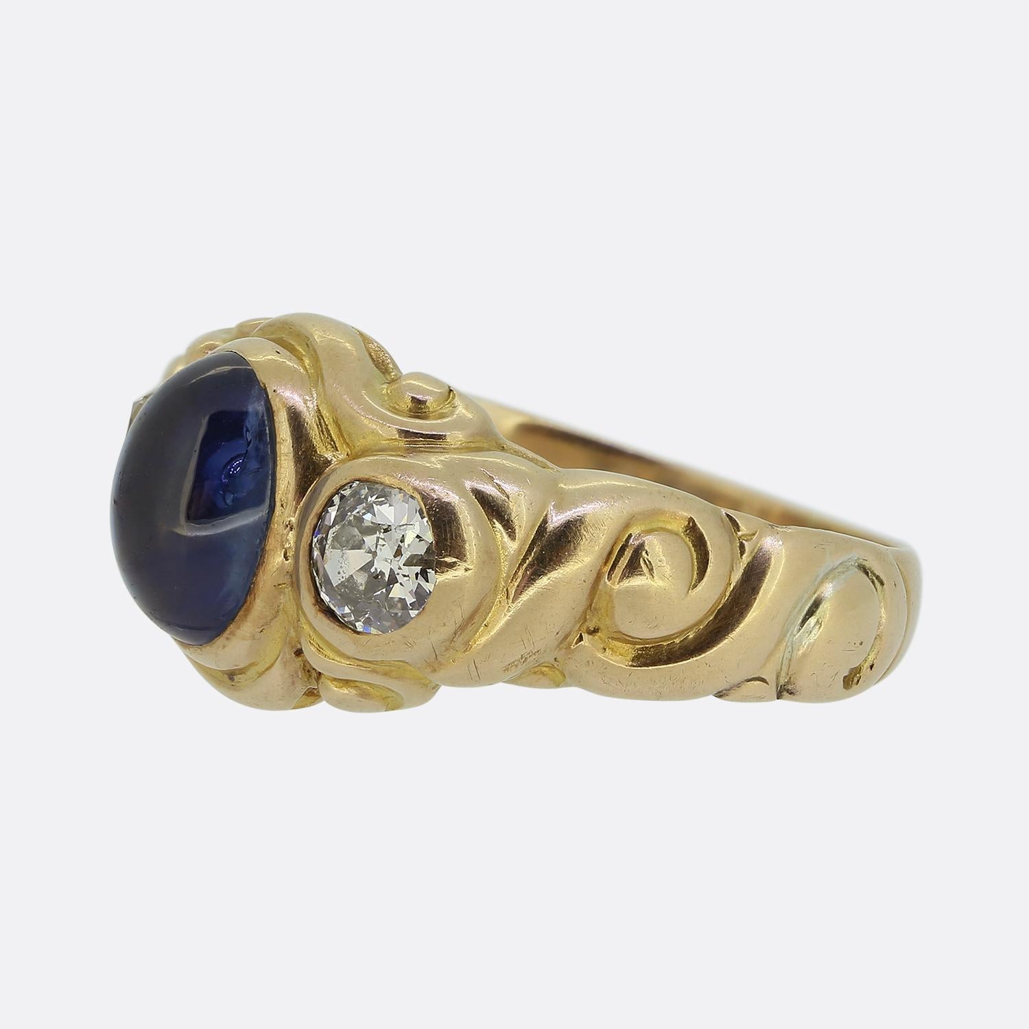 Here we have a distinguished Victorian three-stone ring. Crafted from a rosey 18ct gold, this piece features a centralised medium blue oval shaped cabochon-cut sapphire. This principal stone is then flanked on either side by a single round faceted