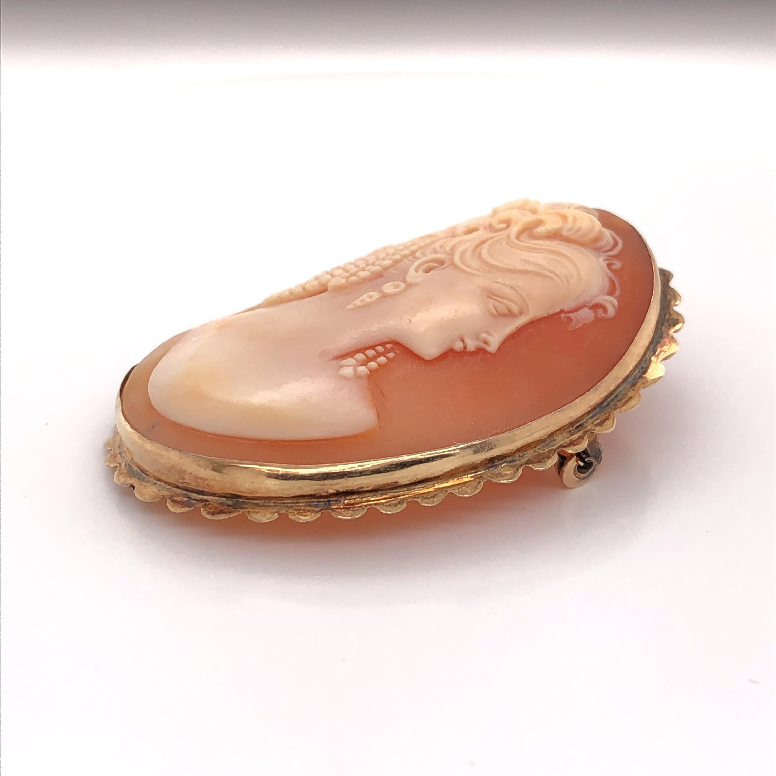 Unusually fine in detail, this hand carved shell cameo celebrates the beauty and soft features of its subject. She sits in a scalloped edge frame of fourteen carat yellow gold. The piece is fitted with a pin and C-closure to be worn as a brooch and