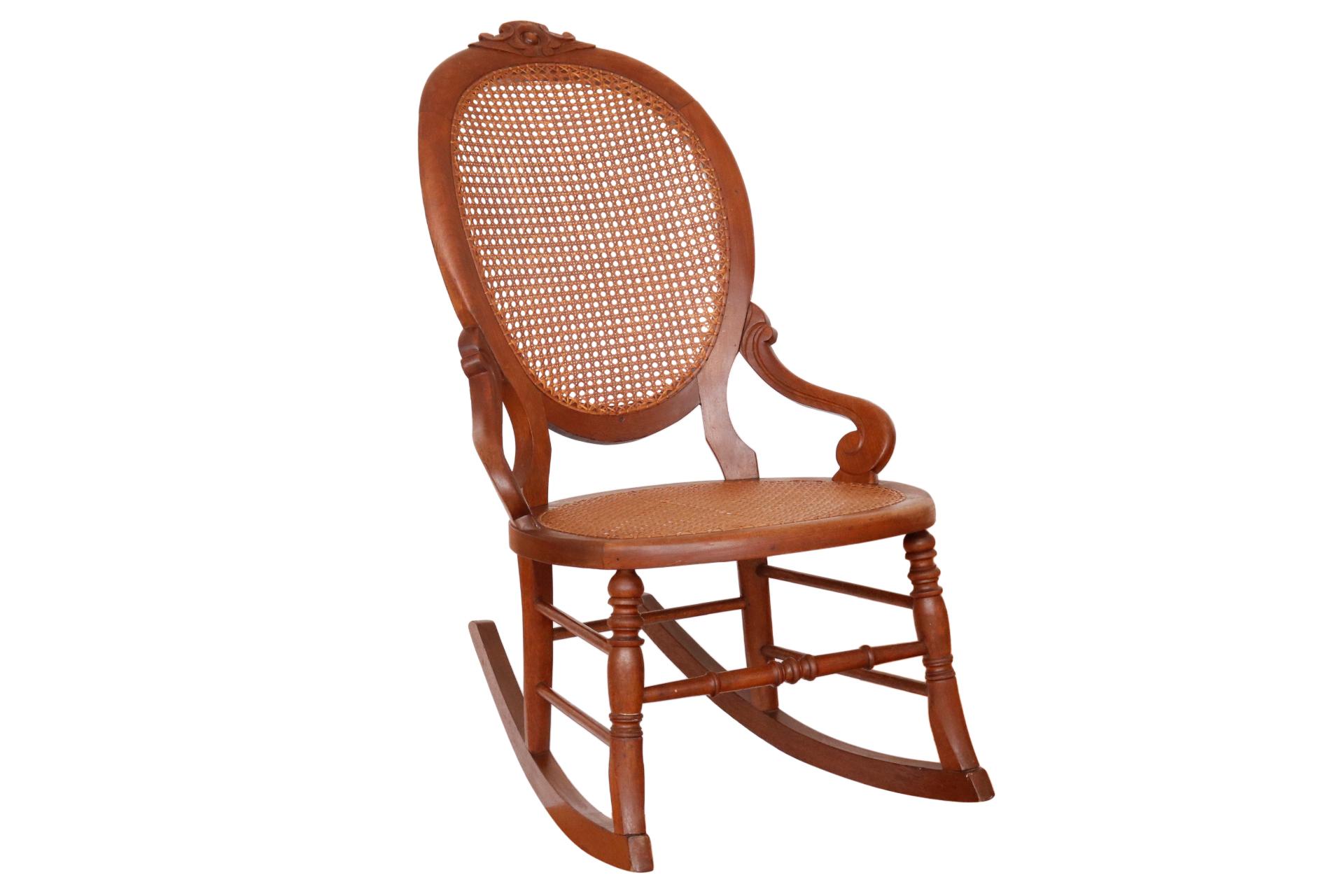 An antique caned rocking chair. The chair has a caned balloon back carved on the crest with a simple flower. Outward flowing arm like supports scroll at the seat. Turned legs are connected with a box stretcher and finish with gently curved rockers.