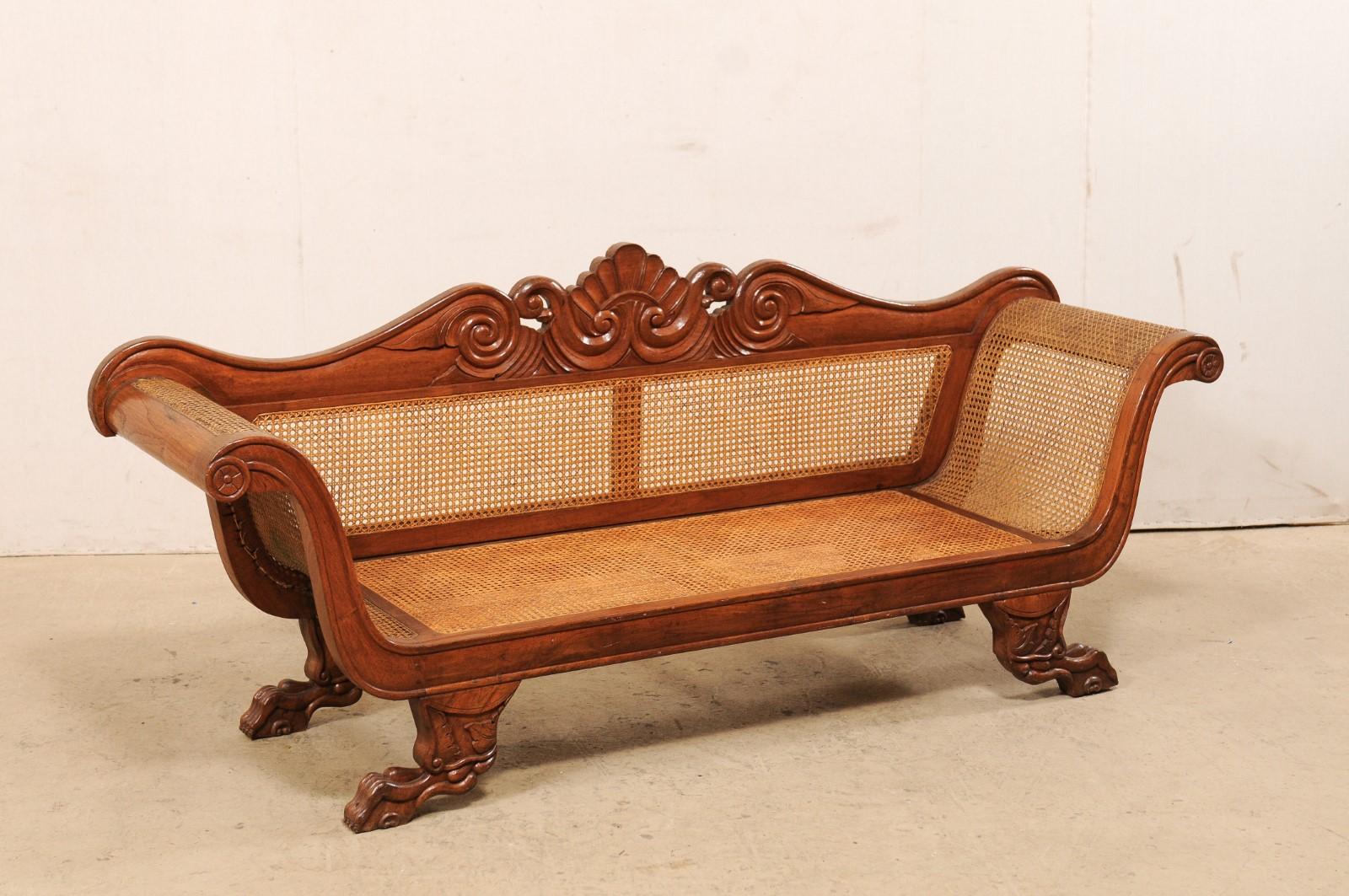 A Caribbean Regency carved-wood and cane sofa from the early 20th century. This antique sofa from Jamaica features a carved crest in shell and wave motif, gracefully curved arms, and is raised on outward facing paw feet, all typical of the Regency