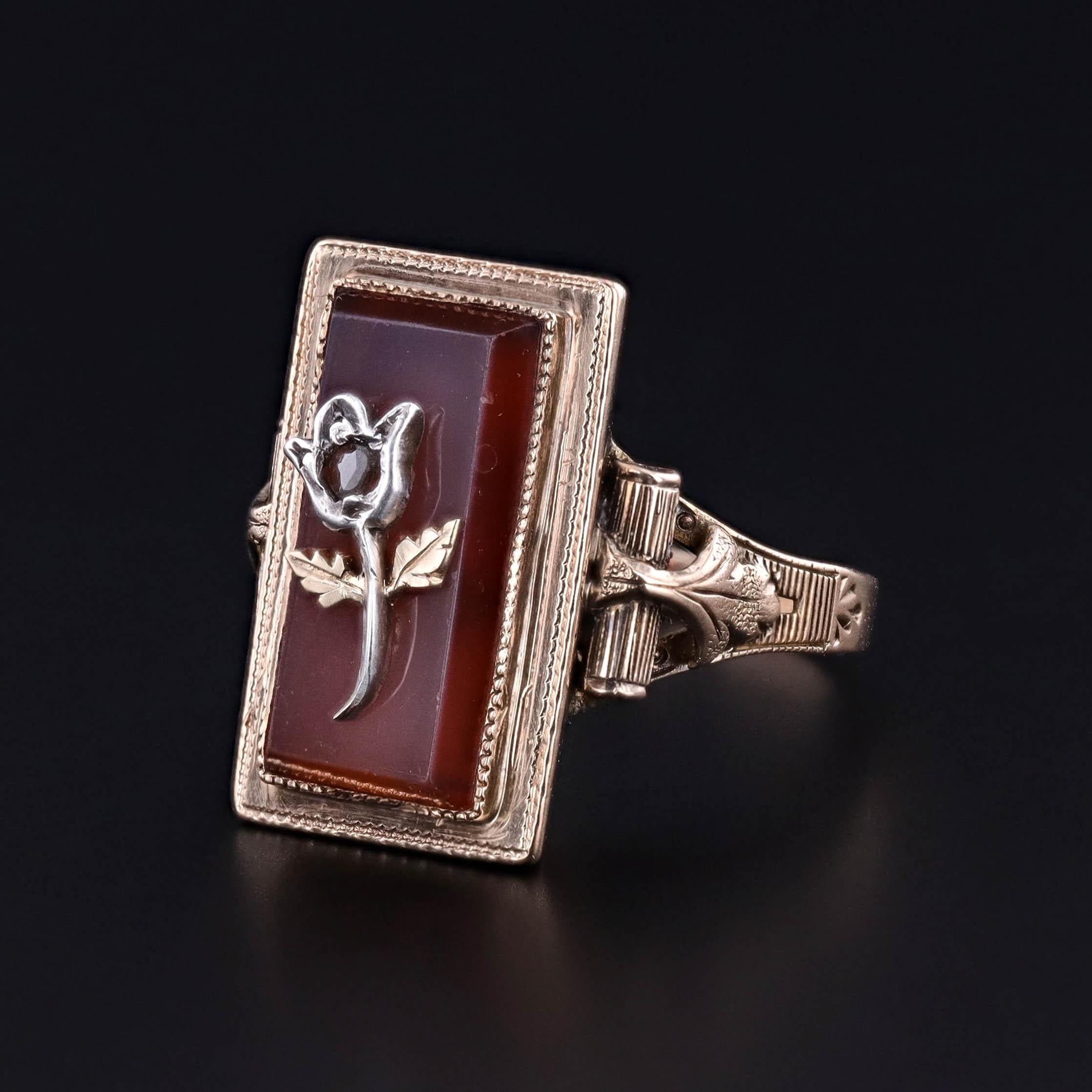 Cette bague ancienne enchanteresse, fabriquée en or 14k, présente une délicate rose sertie de diamants en pleine floraison, posée sur une plaque de cornaline polie. Le contraste frappant entre le doux éclat du diamant et la beauté sombre et terreuse