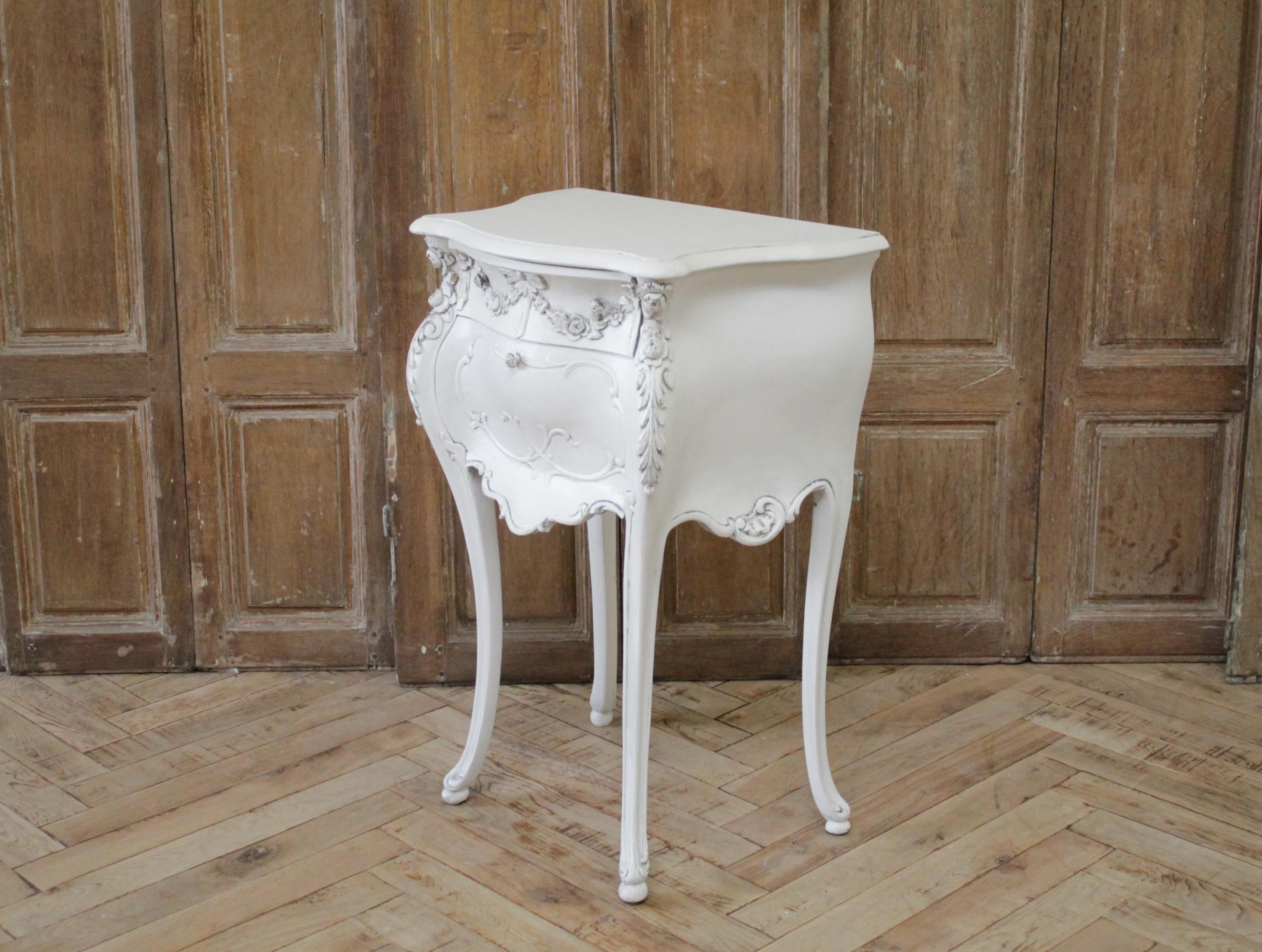 Antique carved and painted nightstand by Louis Bulloni
Painted in a antique white, with subtle distressed edges, and antique patina. This side table has a single drawer and drop down door for storage. 
A beautiful delicate ribbon and rose swag